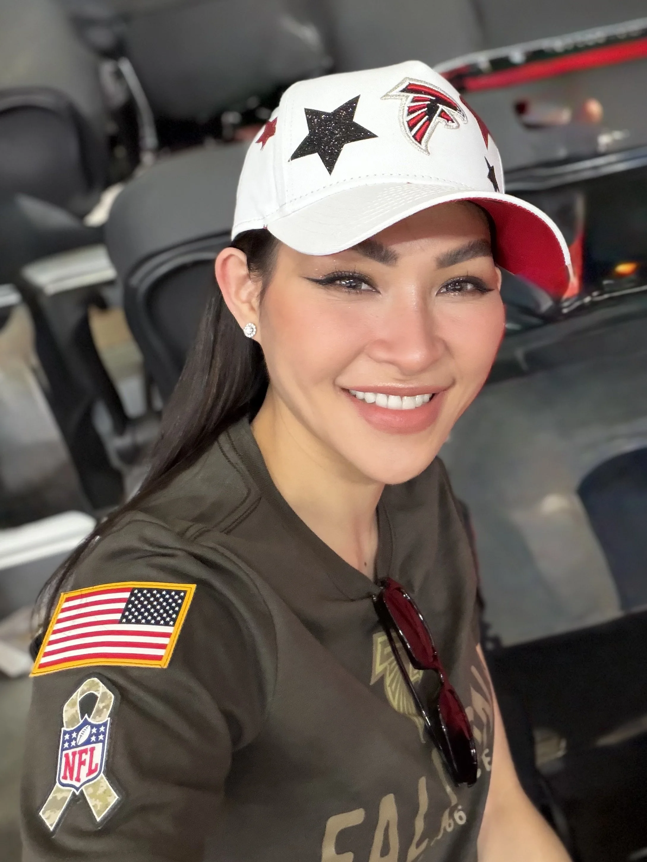 Trang T. Nguyen smiling in an Atlanta Falcons cap and Salute to Service shirt at a football stadium.