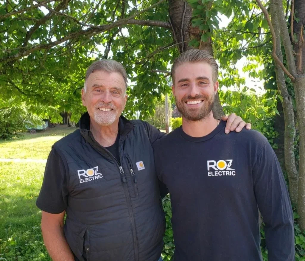 Two smiling men outdoors standing close together with their arms around each other, in front of a tree with green leaves. Both are wearing black shirts or jackets with the 'ROZ Electric' logo.