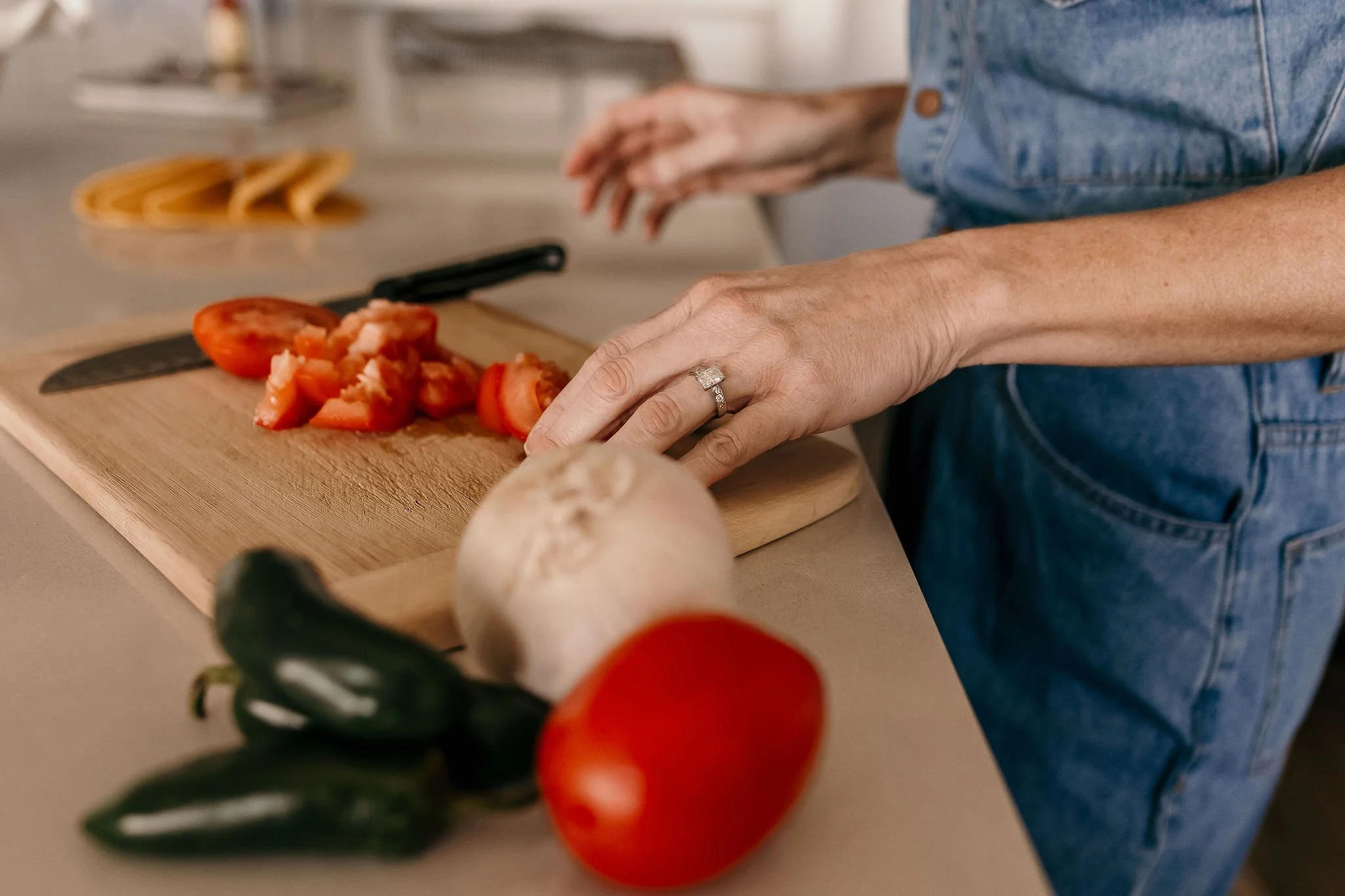 A person making salsa, chopping tomatoes on a wooden cutting board in a kitchen, with fresh vegetables nearby, including jalapenos, a garlic bulb, and a red bell pepper.