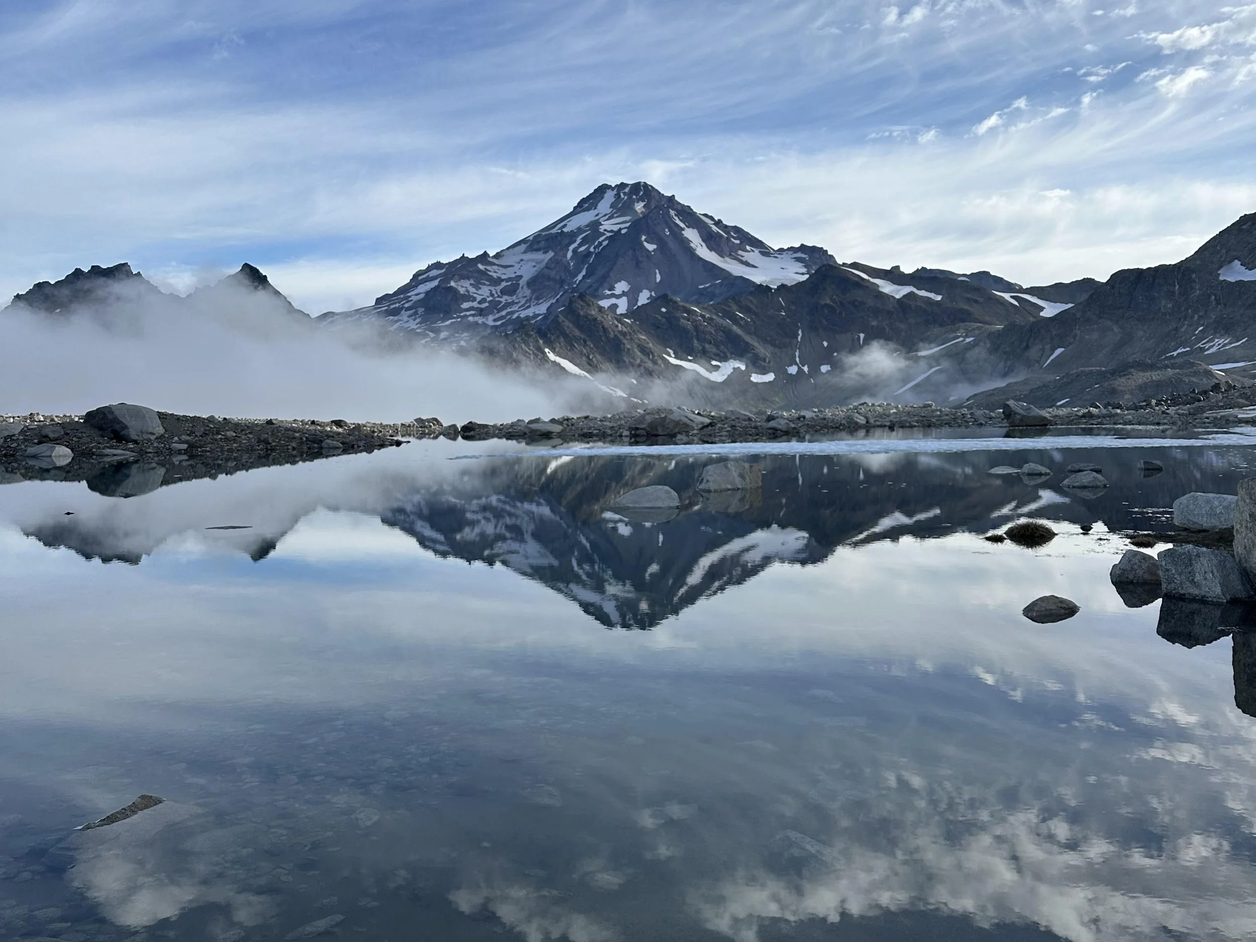 Snow-capped mountain with reflection in a calm lake surrounded by rocks and cloudy sky.