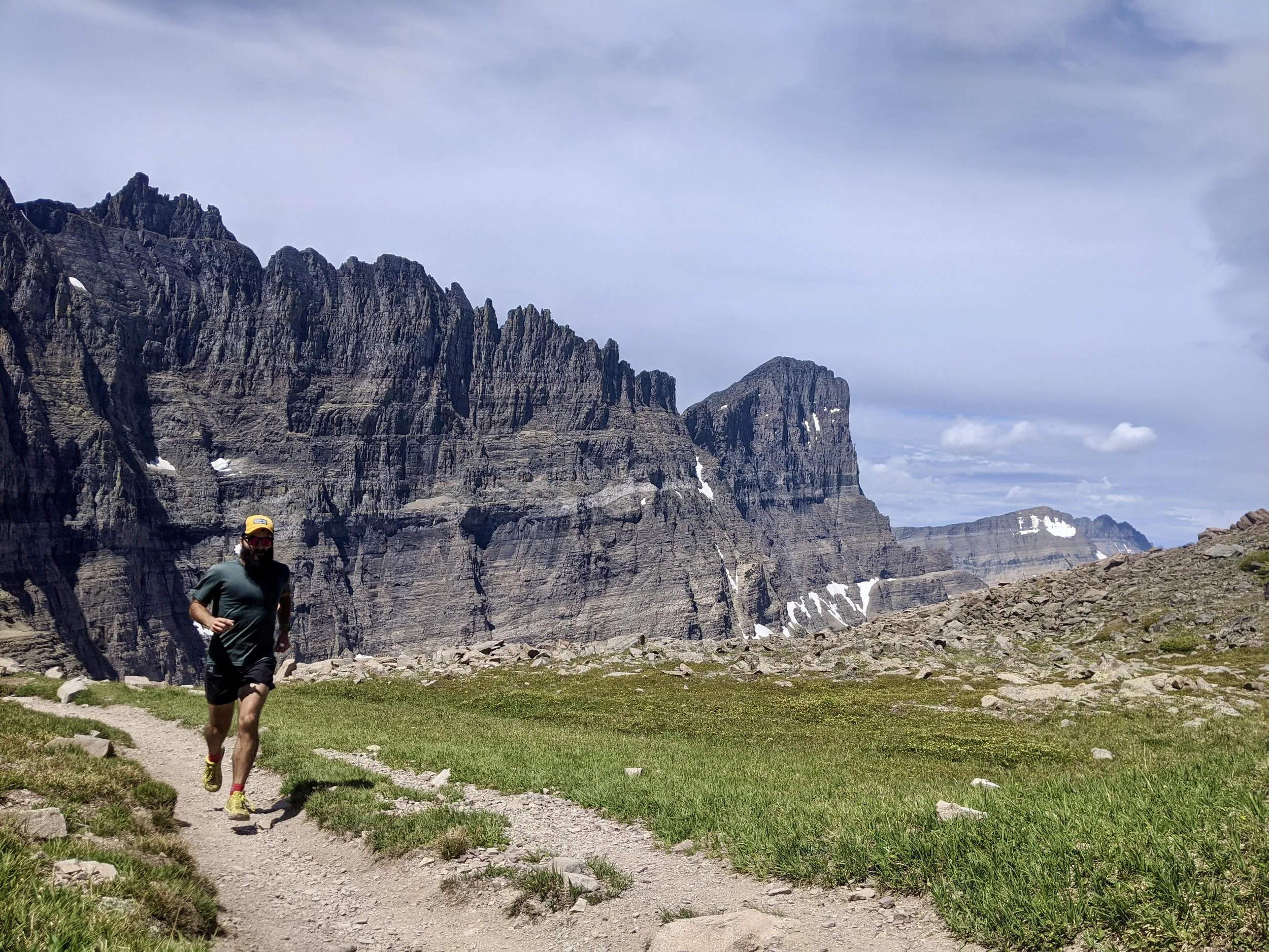 A man running on a dirt trail in a mountainous area with steep rocky cliffs and patches of snow in the background.