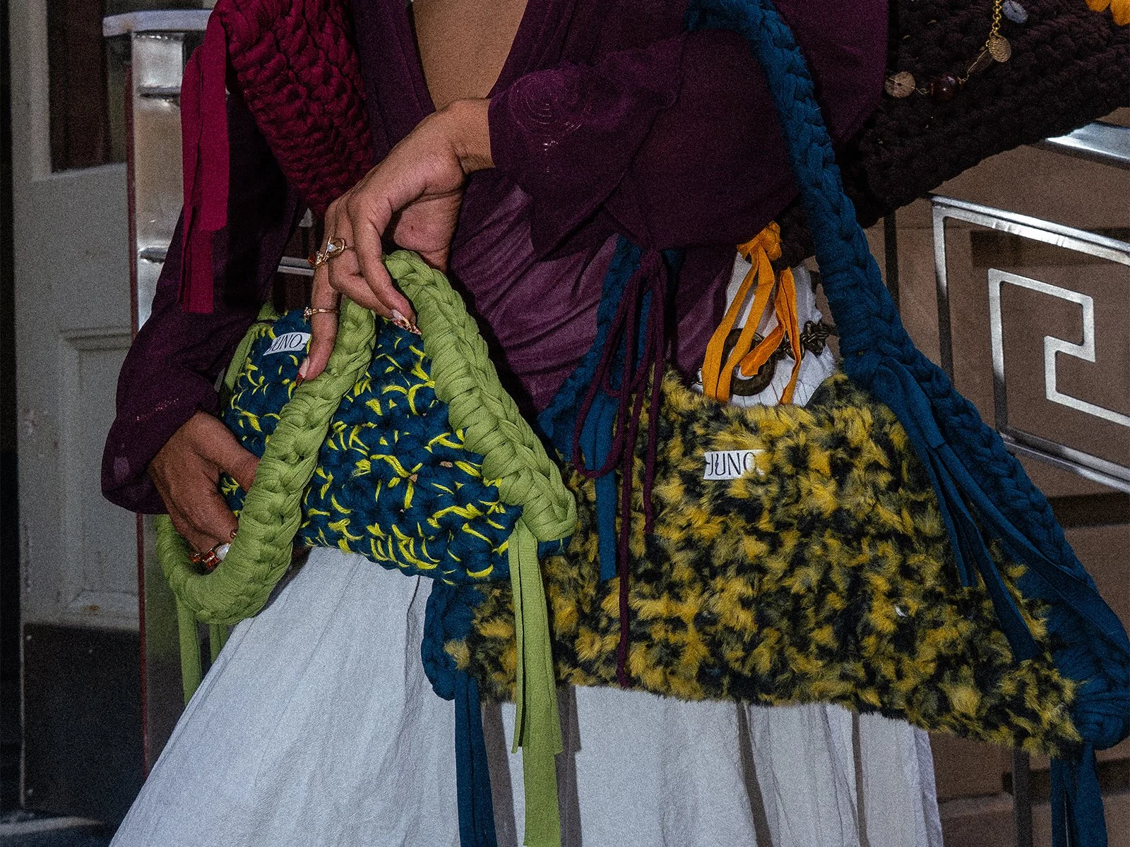 A model holds three of the on of a kind, colourful, Limited JUNO crochet bags in their arms, the city landscape behind them.