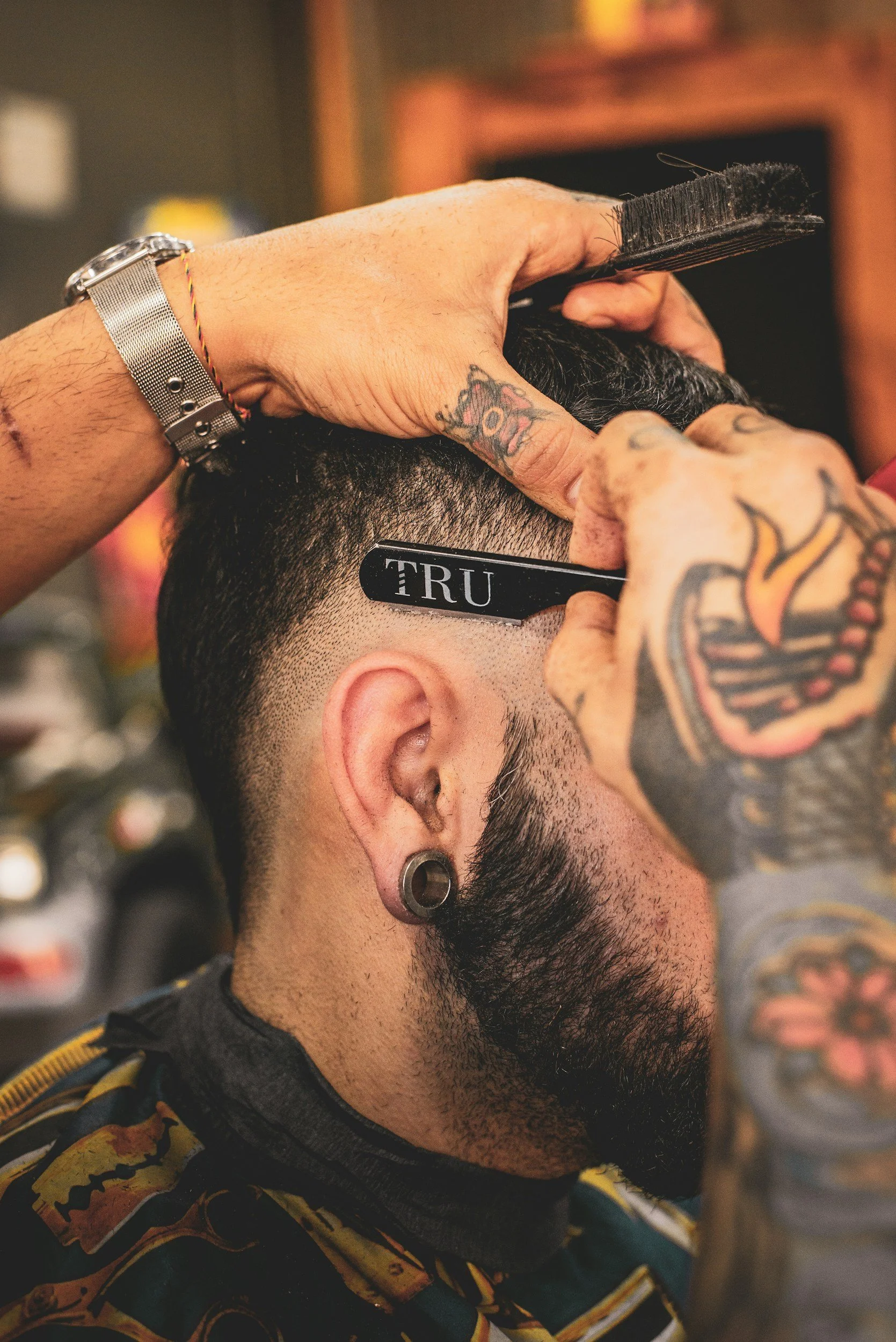 A man gets a haircut at a barbershop, with a barber using a razor to shape his hair. The man has a tattoo on his hand, earrings, and a watch, and is wearing a colorful patterned shirt.