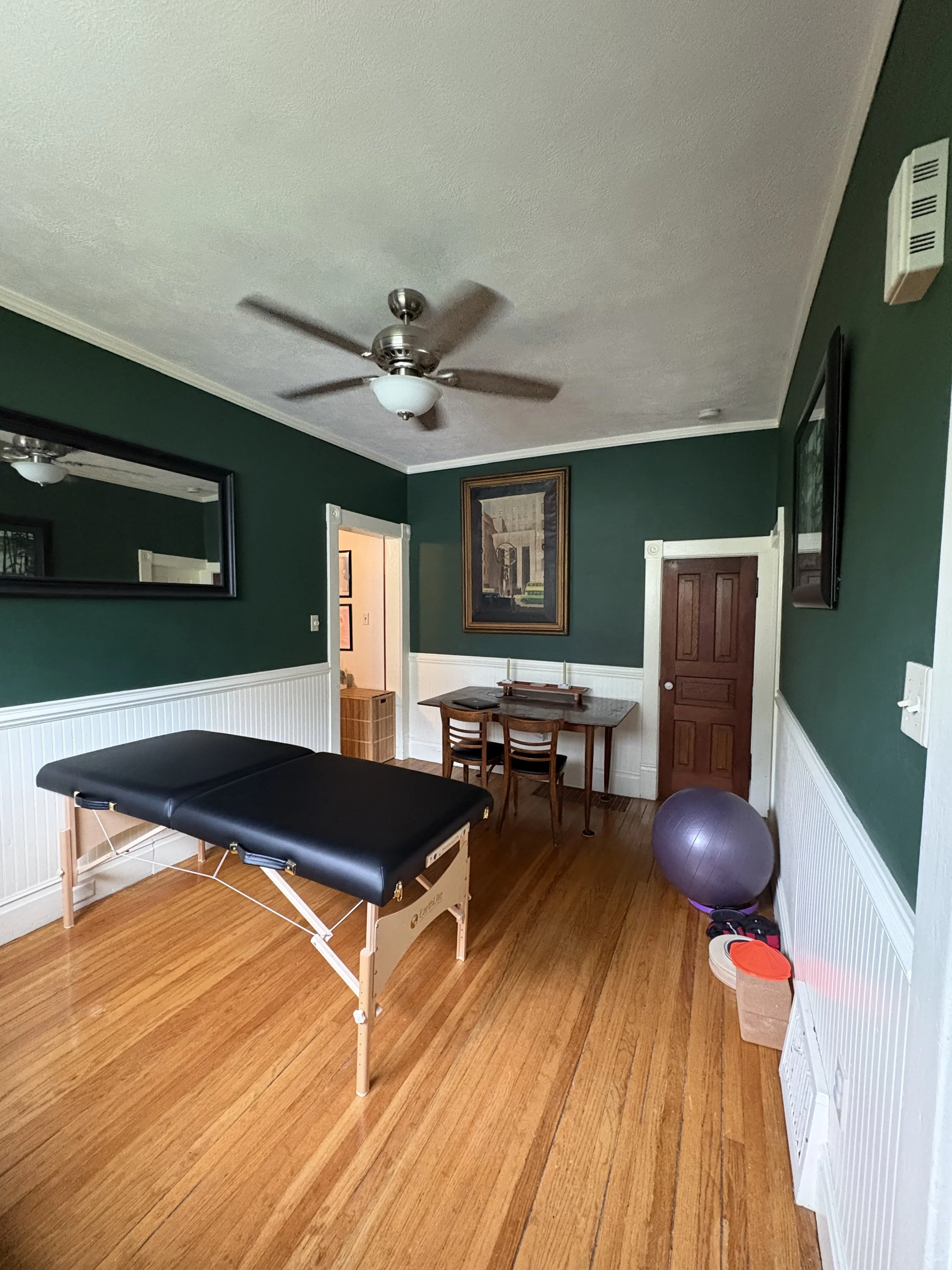 Massage table set up in a green room with wooden floors for concierge physical therapy services, along with a wooden table, swiss ball, cones, and fan on ceiling