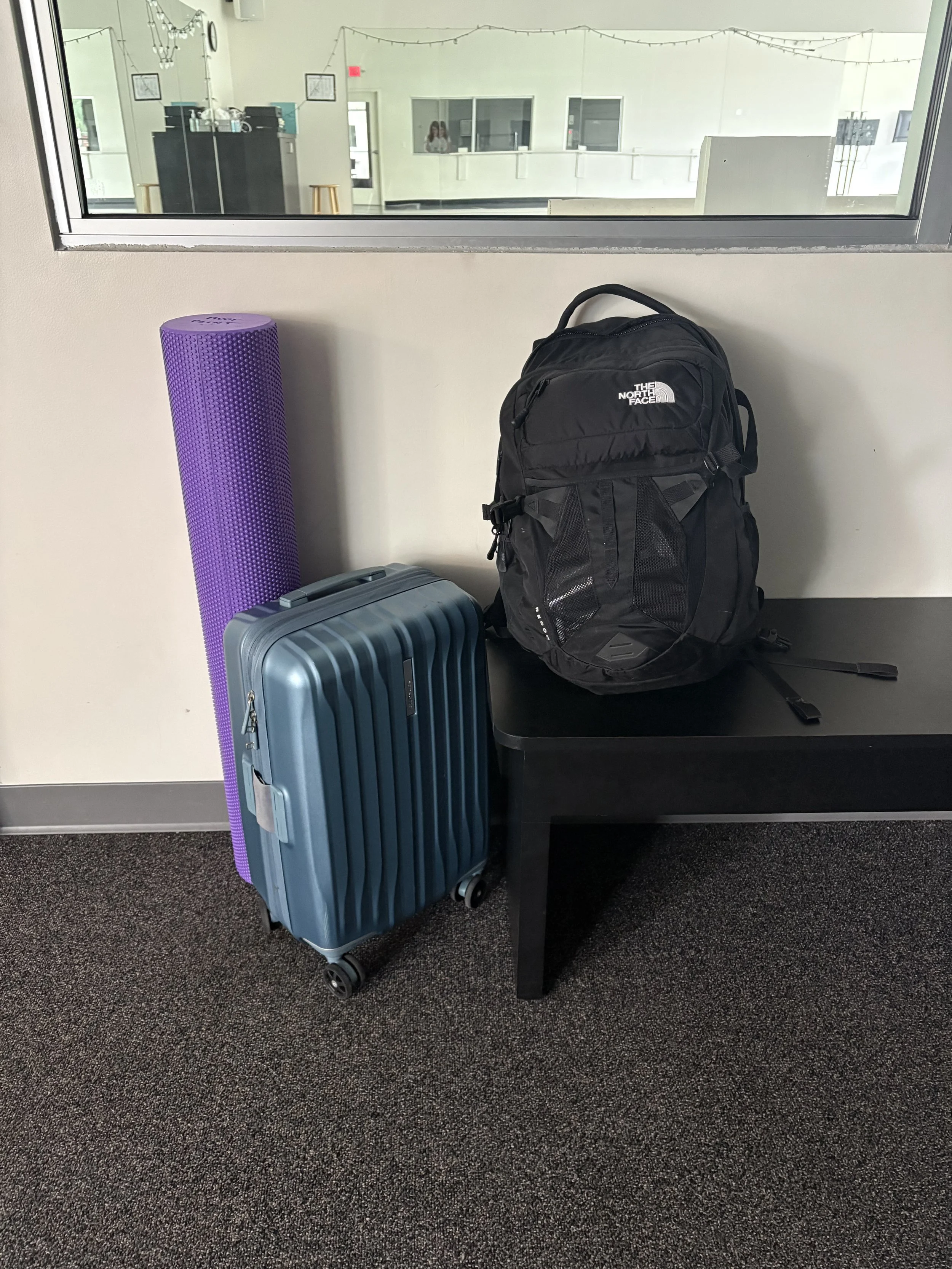 Foam roll, rolling luggage, and black backpack filled with physical therapy supplies for mobile treatment at a dance studio, with white walls and a ballet barre in the background.