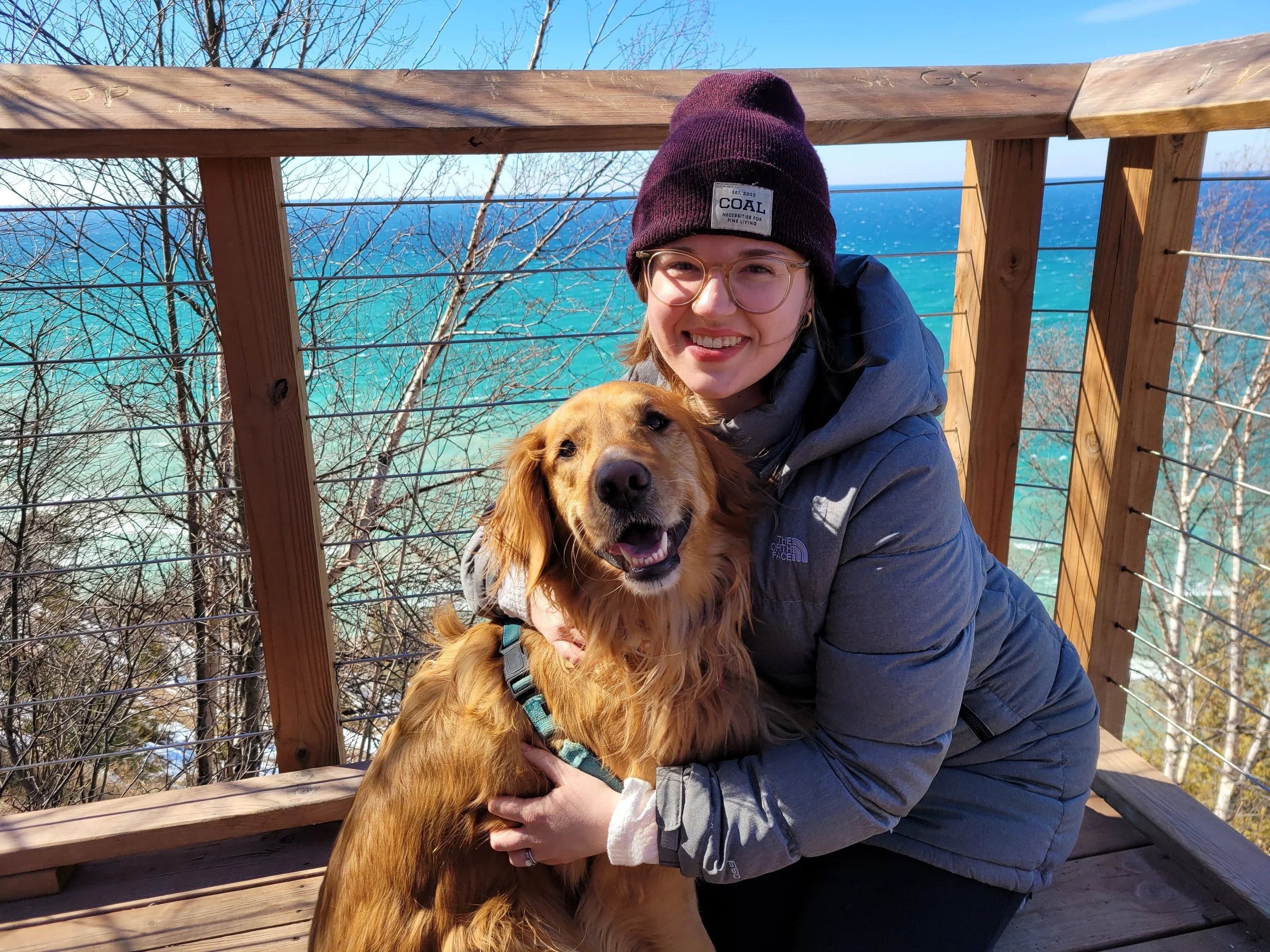 A woman in a gray jacket and purple beanie with glasses, smiling and hugging a golden retriever on a wooden deck with ocean and bare trees in the background.