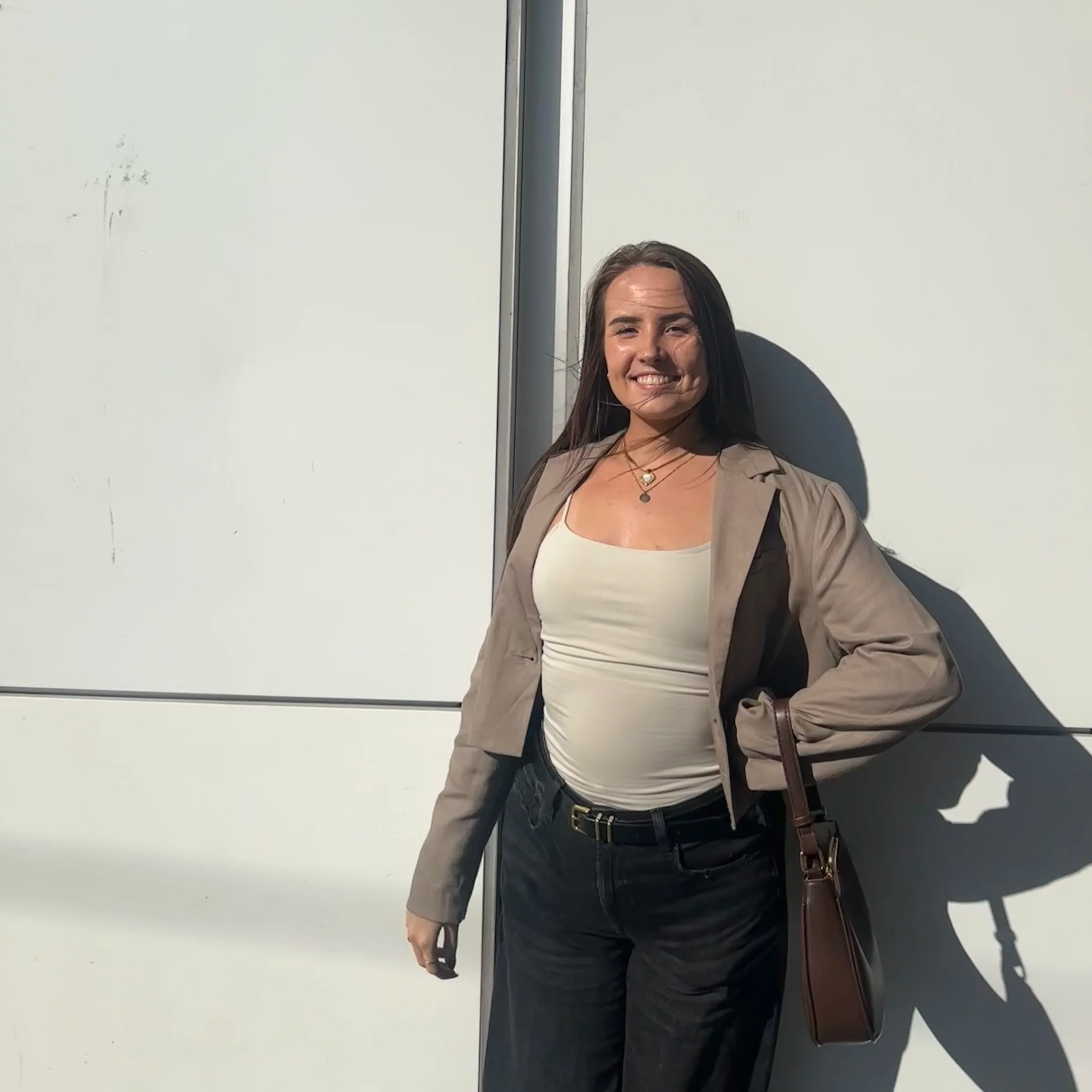 A woman standing against a white wall, smiling, wearing a beige jacket, a white top, black pants, and holding a brown purse.
