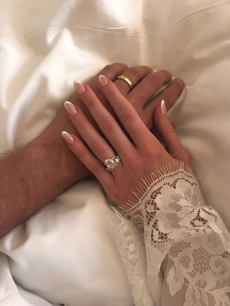 Close-up of a couple's hands with wedding rings on a white satin fabric, with one hand wearing a lace sleeve. Bronzed hands showing off the custom airbrush tan they got prior to the wedding.