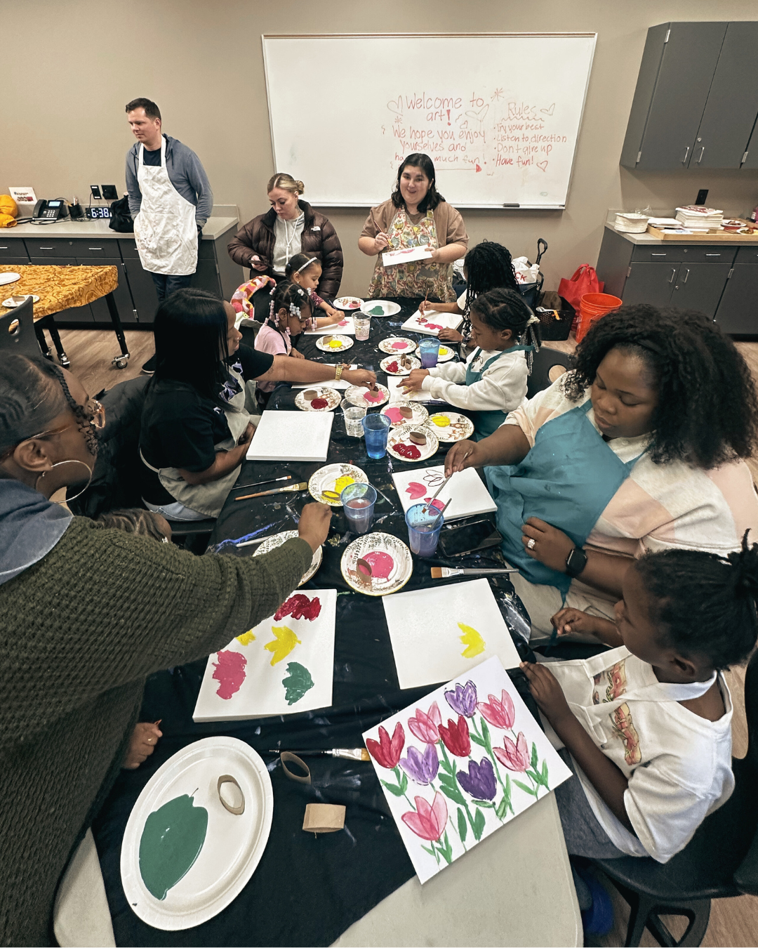 Children and adults are painting flowers on paper at a classroom art activity at Family First Community Center