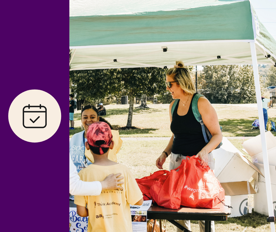 A woman with sunglasses, light brown hair, and a green backpack talking to two children at an outdoor event under a white tent. The children, a boy in a yellow t-shirt and a girl in a gray sweatshirt, are facing the woman. The woman is holding a red bag, and there are boxes on the table. The background shows trees and other people at the event.