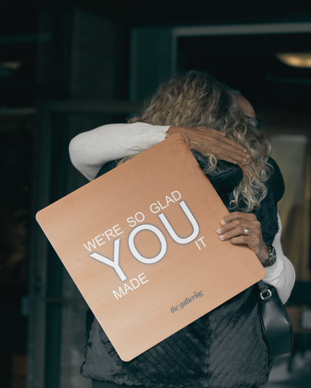 Two women hugging, one holding a sign that says "We're so glad you made it" in a warm tone, indoor setting.