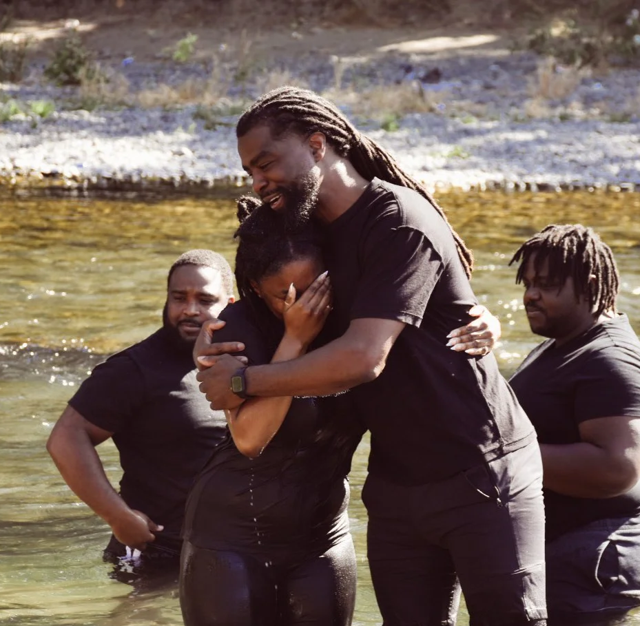 A group of four people standing in a river, with two hugging and two watching, during a sunny day.