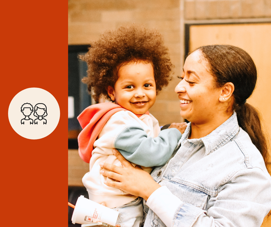 A smiling woman holding a young girl with curly hair, both appearing happy indoors.