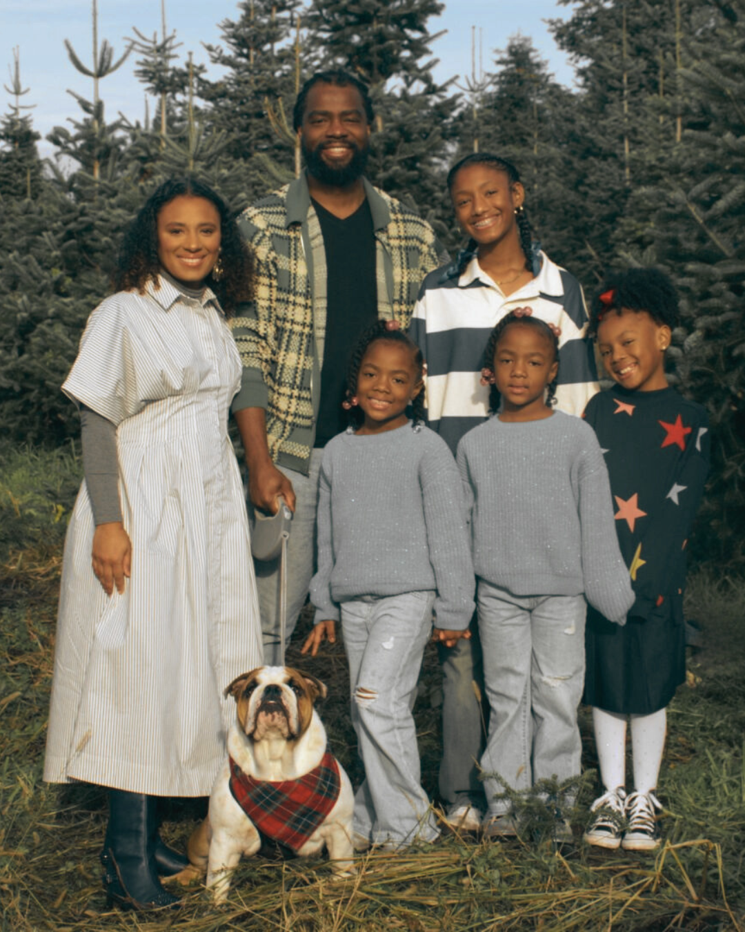 A family and their dog posing outdoors among Christmas trees, smiling at the camera.