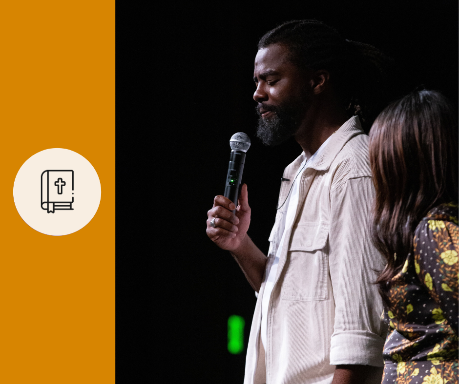 A man with long hair and a beard holding a microphone in his right hand speaking or praying with eyes closed, standing beside a woman on a dark stage.