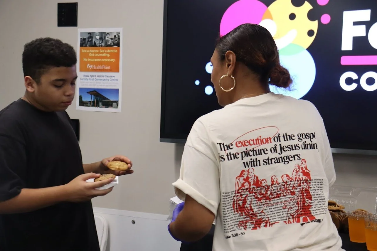 A woman with hoop earrings and a white shirt with a red and black graphic and text is serving cookies to a young boy in a black shirt. The background features a large screen with colorful circles and part of a sign on the wall about healthcare services.