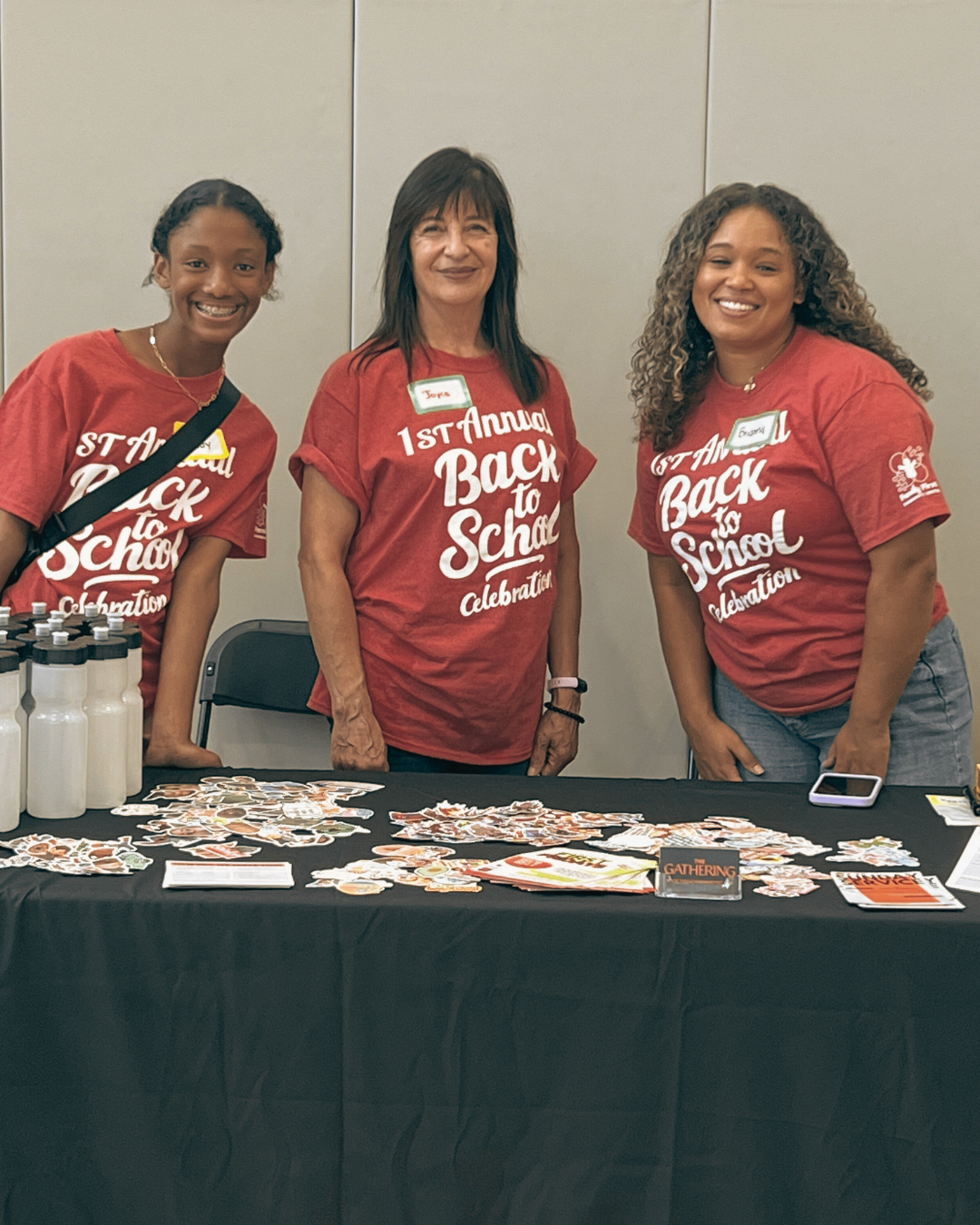 Three women wearing red shirts that say '1st Annual Back to School Celebration' standing behind a table with stickers, pamphlets, and water bottles, smiling at the camera.