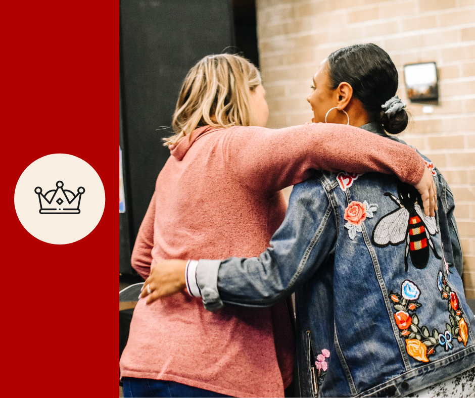 Two women hugging each other, one in a pink coat and the other in a denim jacket with floral and bee embroidery, in front of a brick wall.