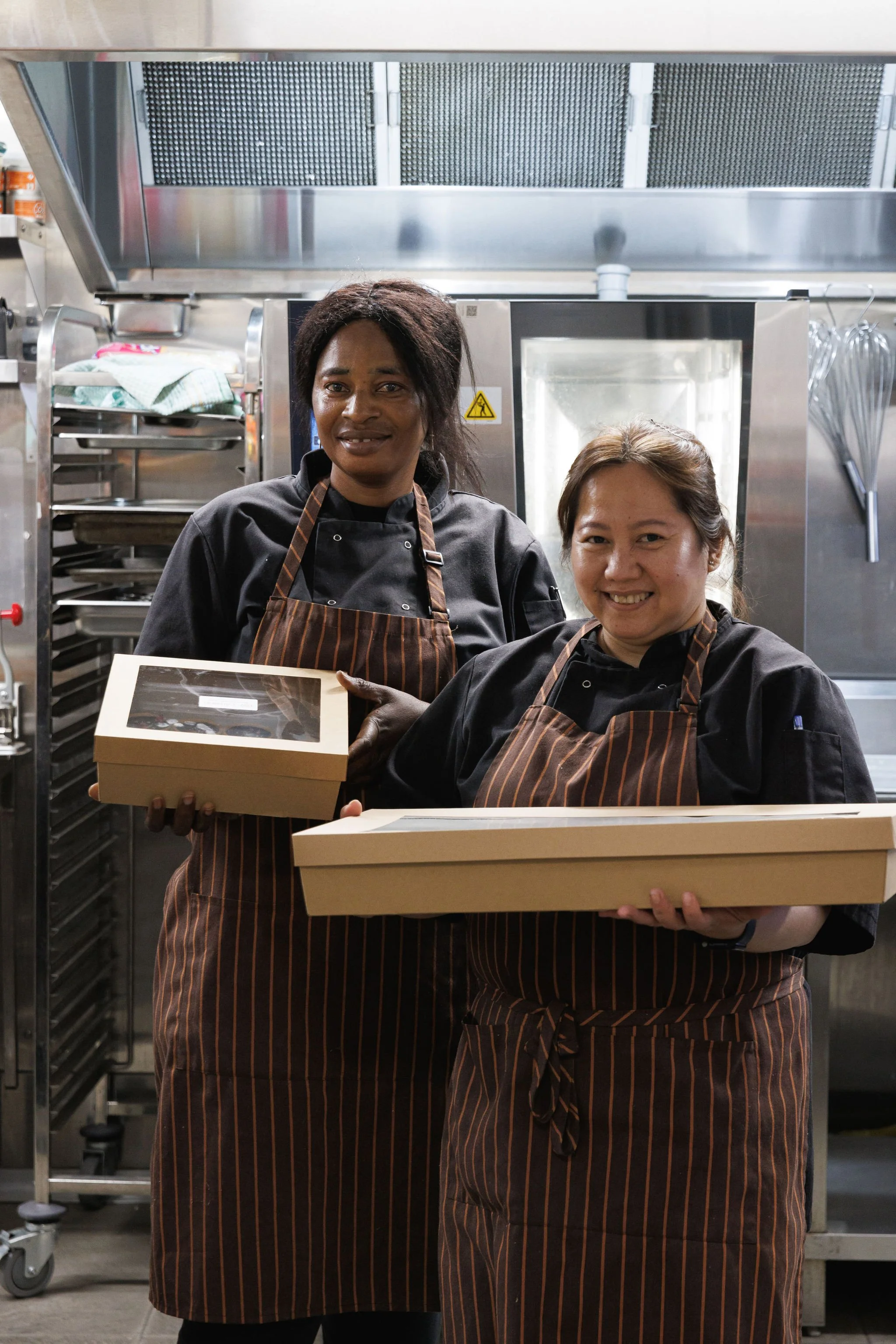 Two women in chef uniforms and striped aprons holding bakery boxes in a commercial kitchen.
