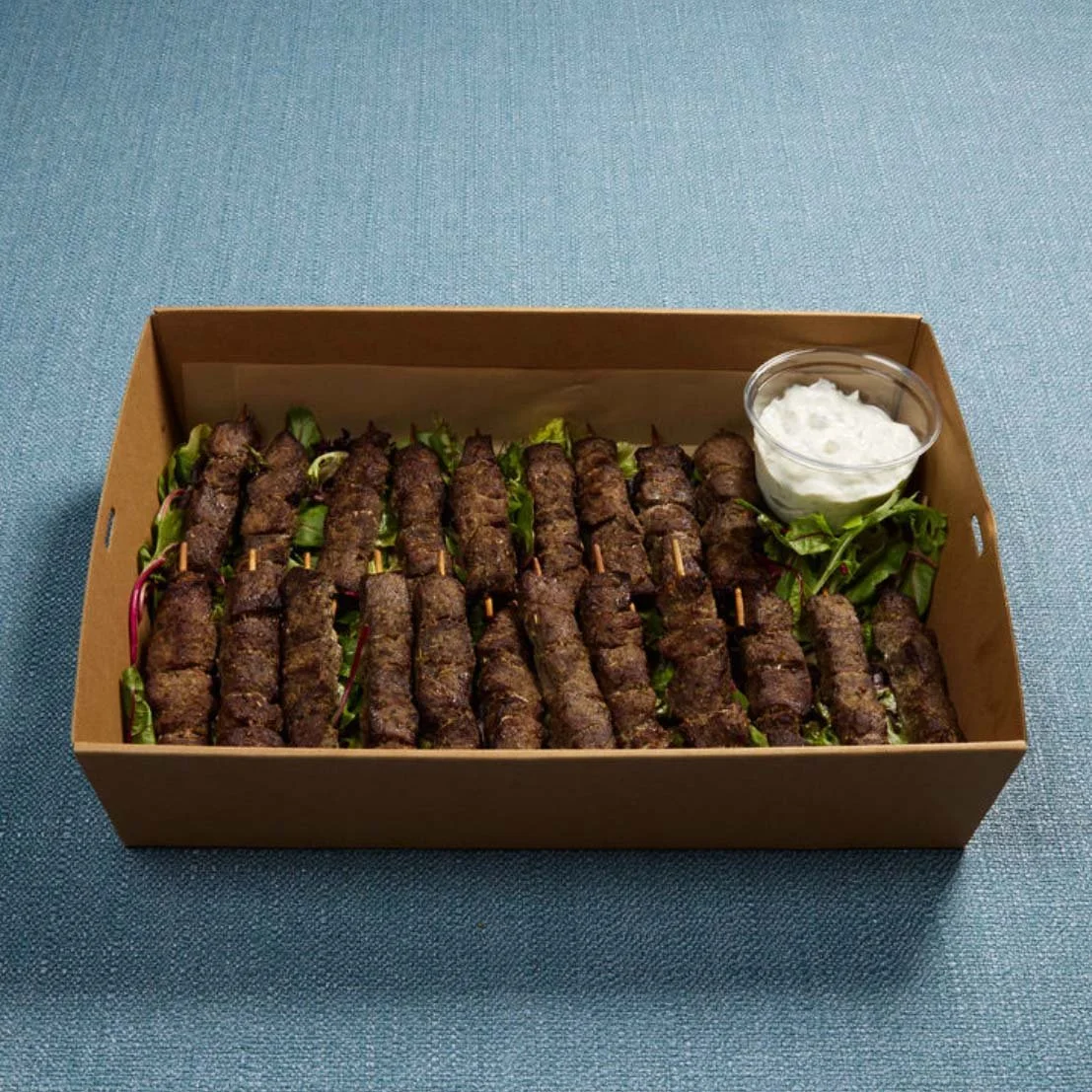 A tray containing grilled meat skewers with leafy greens and a side of white dipping sauce.