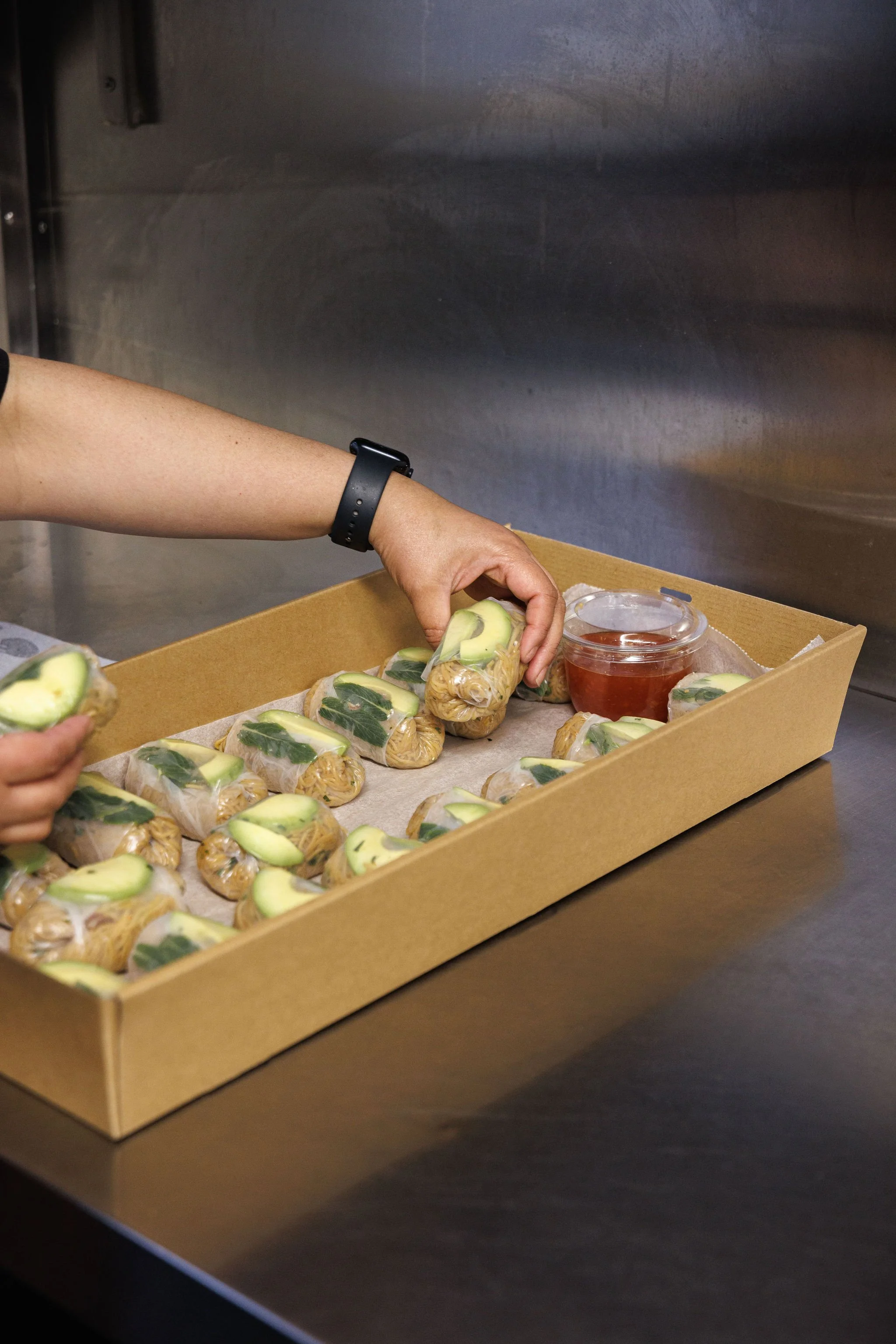 A person packing multiple individual servings of a dish with avocado slices, using a disposable container, with a small cup of sauce or soup nearby.