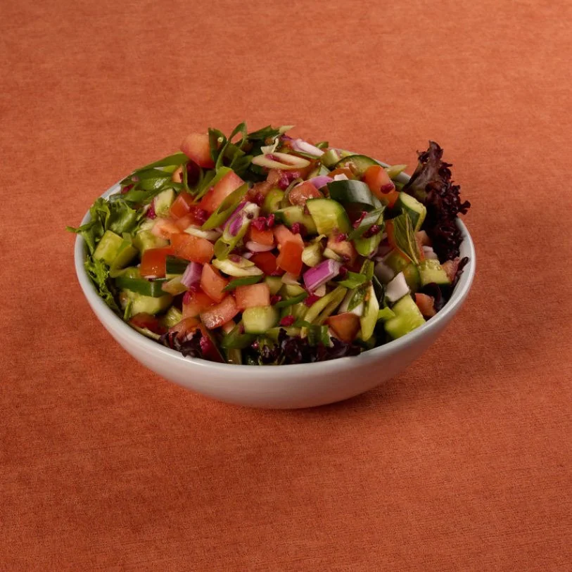 Bowl of chopped vegetable salad containing cucumbers, tomatoes, red onions, green onions, and possibly other fresh vegetables, placed on an orange surface.