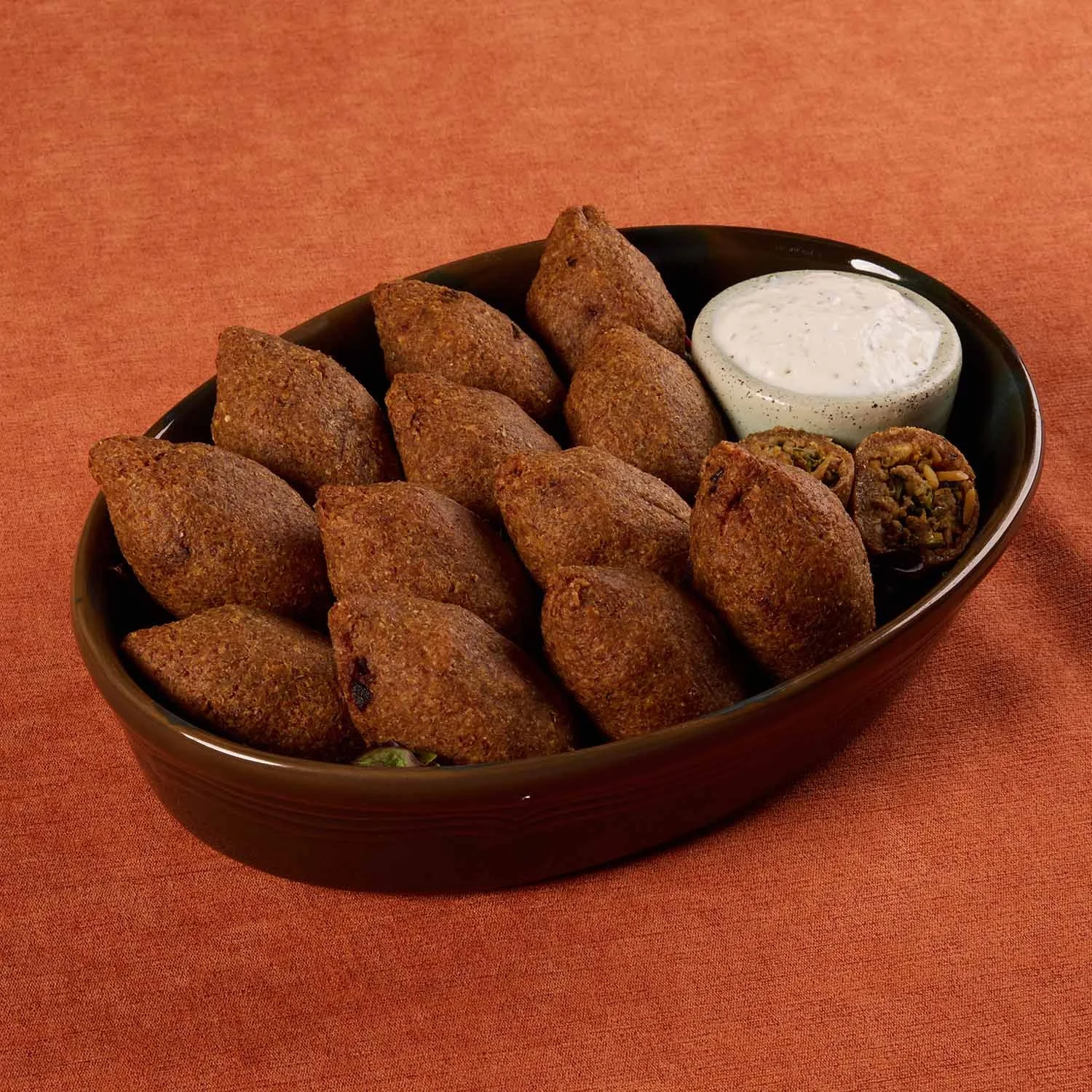 A black oval dish filled with fried falafel balls, a small cup of white dipping sauce, and two pieces of stuffed grape leaves on an orange tablecloth.