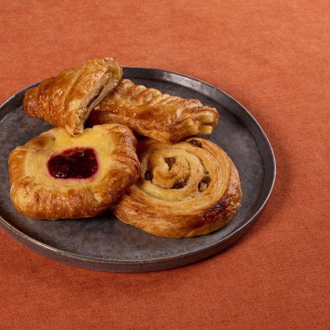 Plate with three different pastries, including a Danish with raspberry jam, a croissant, and a cinnamon roll on an orange table.