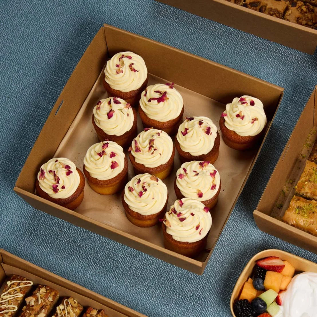 A box of cupcakes with white frosting and pink flower petals on top, arranged in a cardboard box on a blue textured surface.