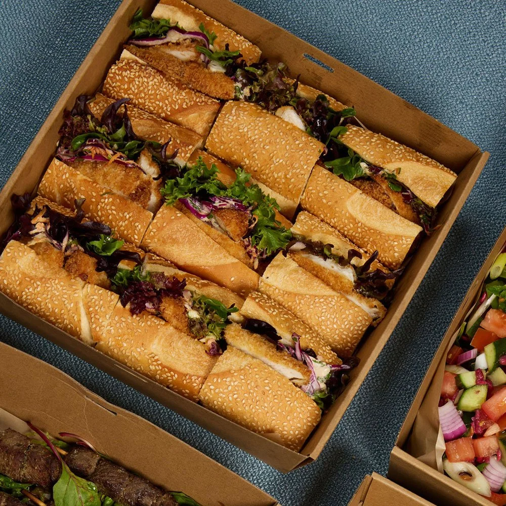 A box of cut sandwich baguettes with fried chicken, lettuce, and purple/red onion.