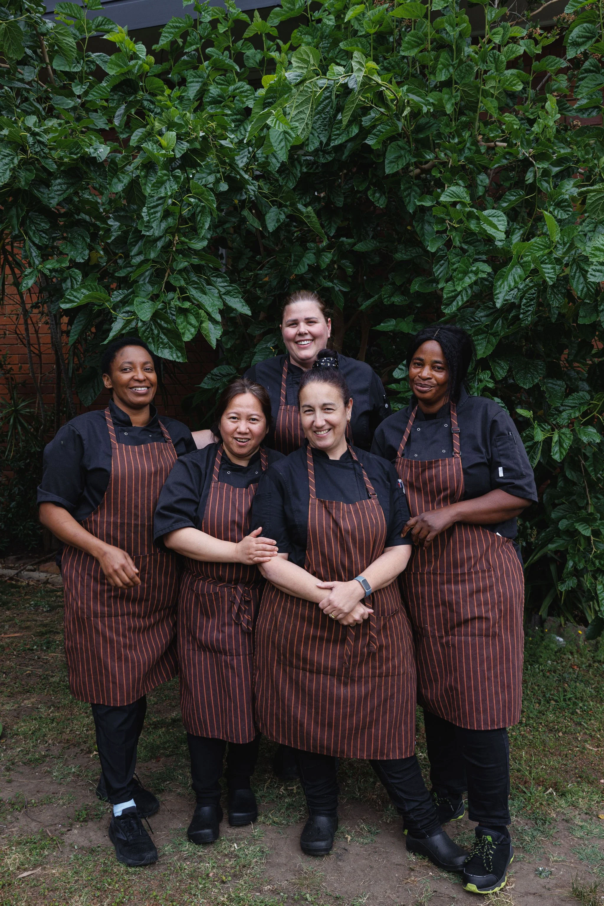Group of five women chefs smiling outdoors, standing in front of leafy green plants, wearing black chef uniforms and striped aprons.
