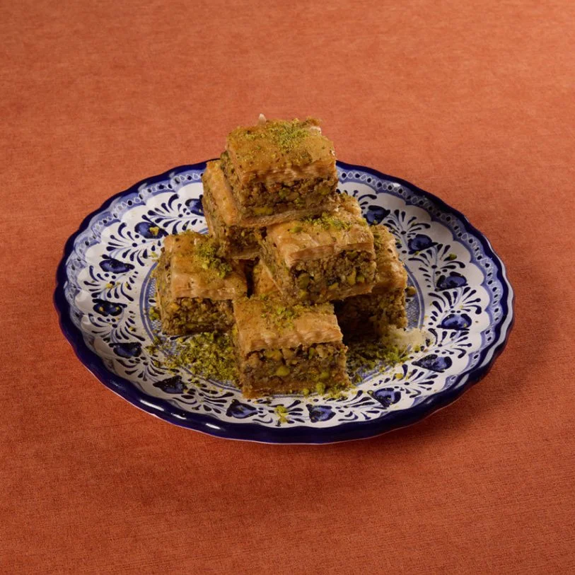 Plate of stacked pistachio baklava with flaky layers and crushed pistachios on top, on a decorative blue and white plate.