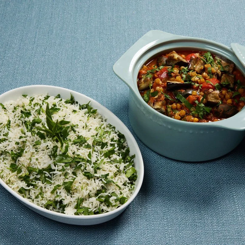 A white oval dish of cooked white rice with chopped green herbs and a blue pot of vegetable stew with eggplant, chickpeas, and tomatoes.