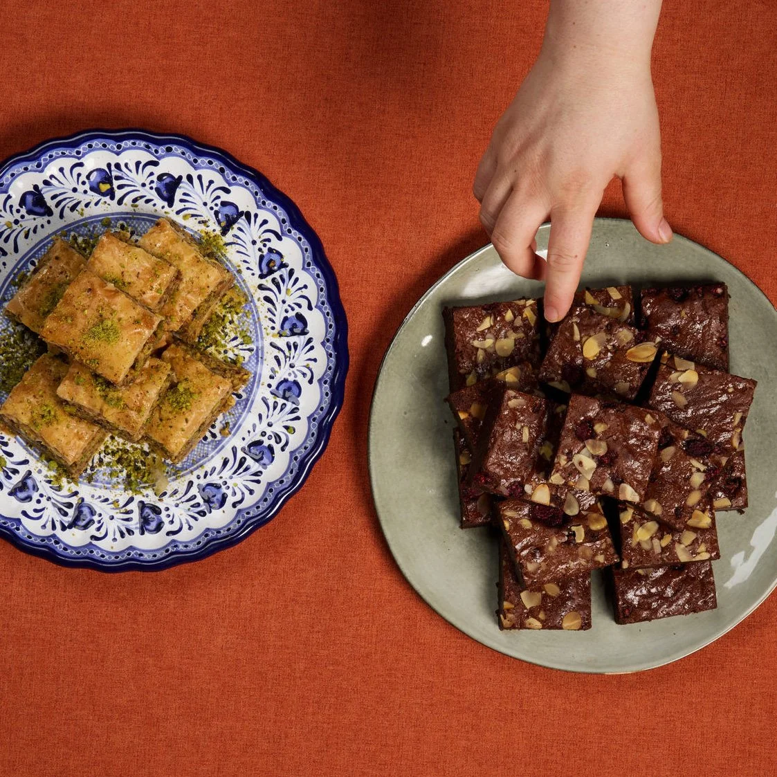 A hand reaching for chocolate almond fudge squares on a grey plate, with pistachillo baklava on a blue and white patterned plate on a red tablecloth.