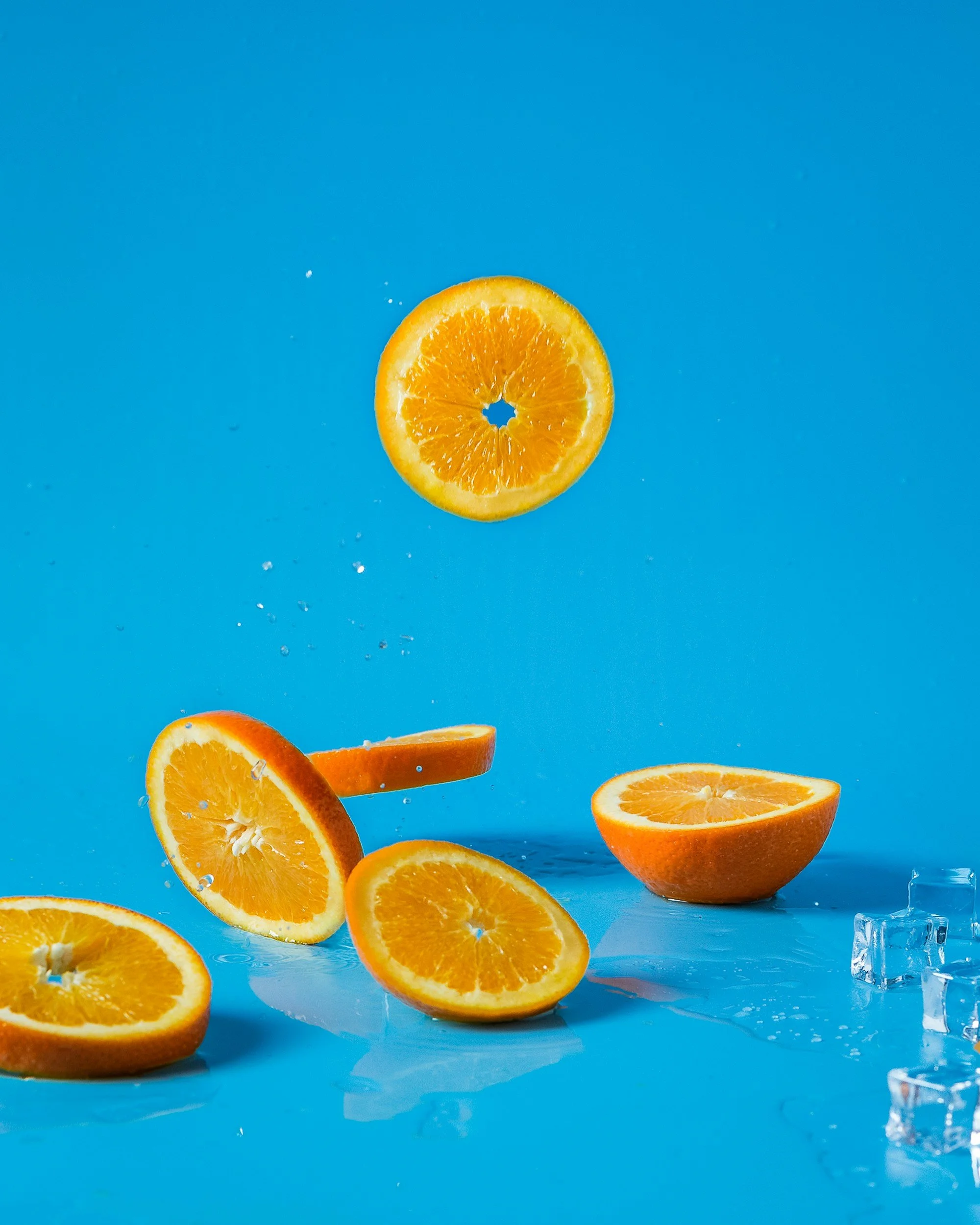Sliced oranges falling into water with ice cubes on a blue background.