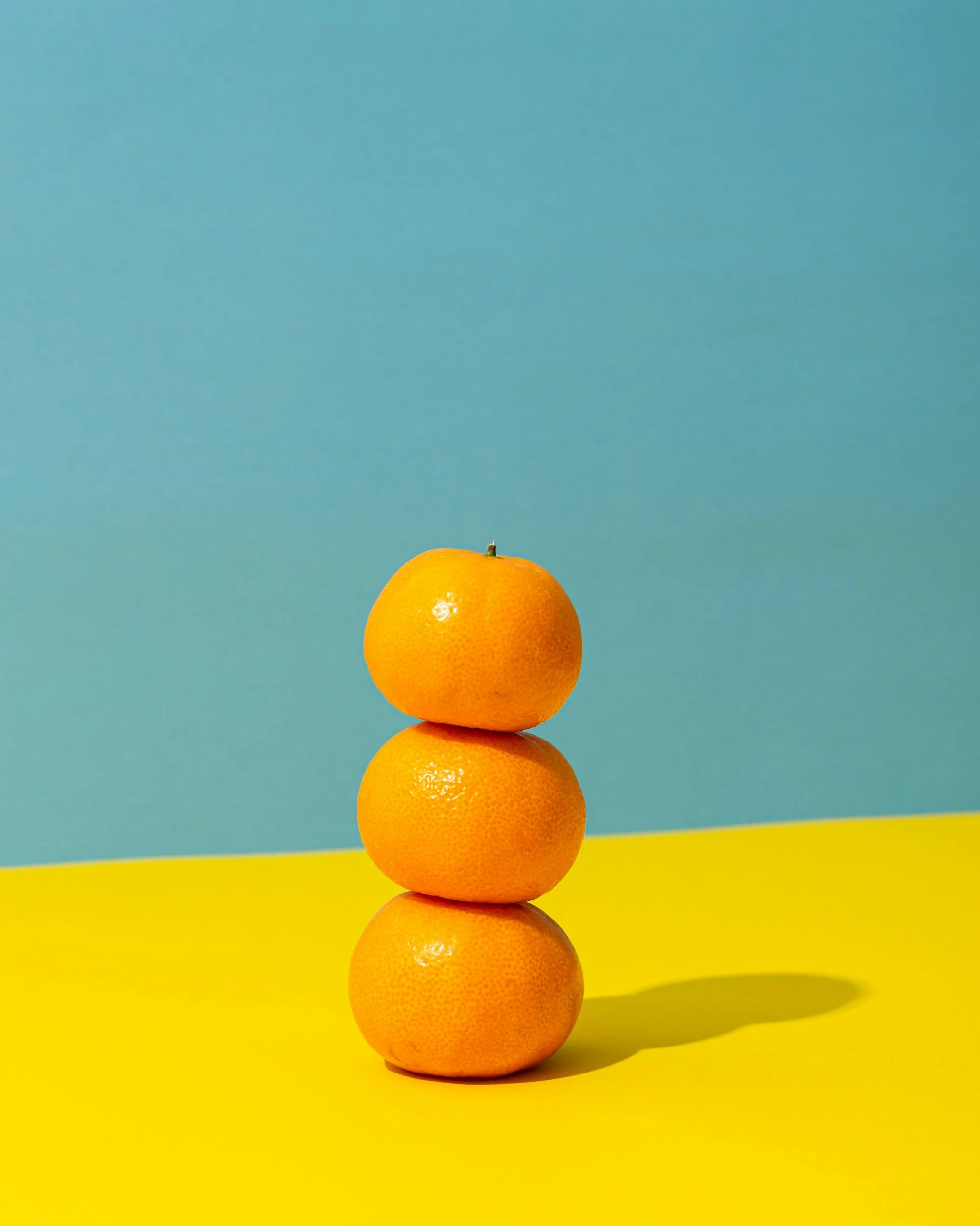 Three oranges stacked vertically on a yellow surface with a blue background.