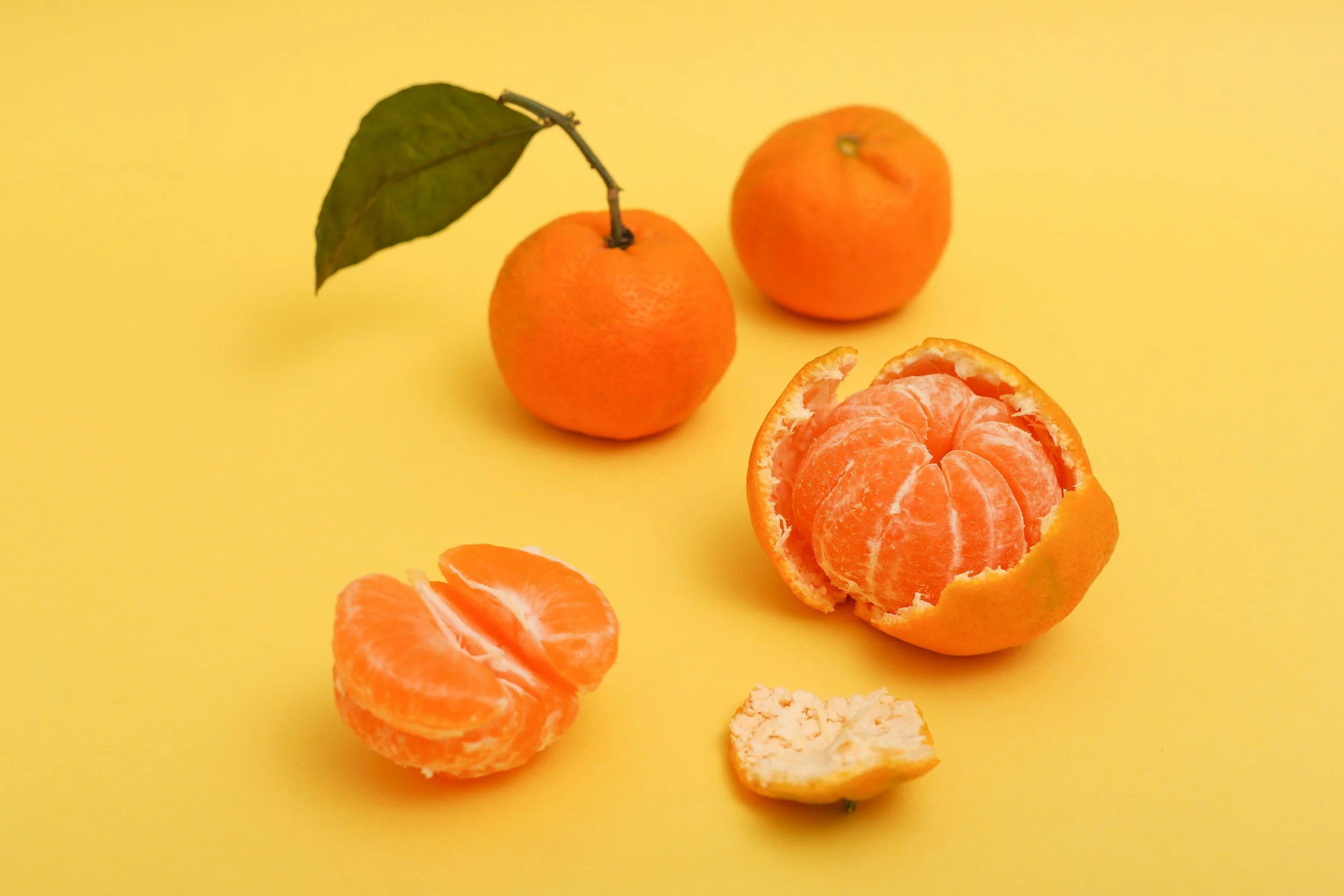 Two whole oranges, one with a leaf on the stem, and a peeled orange with segments and a piece of peel on a yellow background.