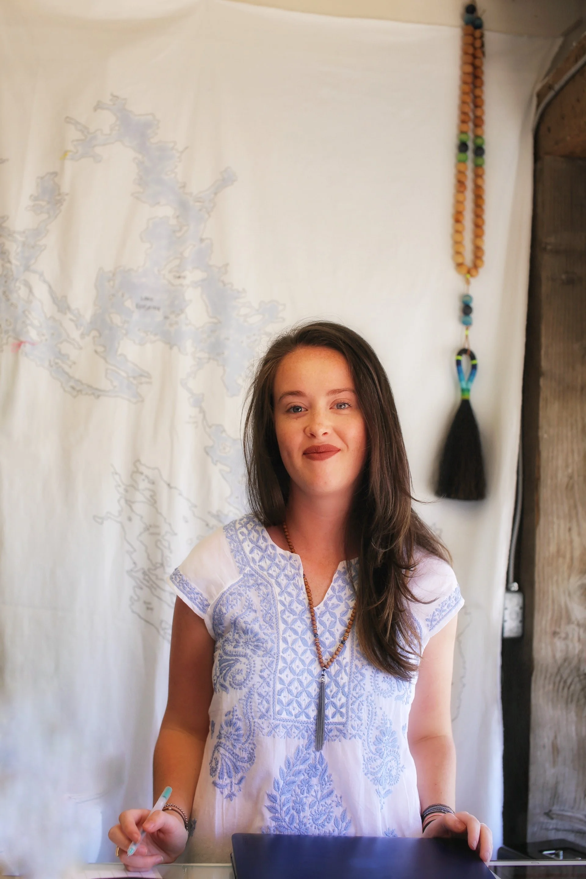 A woman with long dark hair wears a white embroidered top and a beaded necklace. She is standing in front of a white background with a faint map outline and a beaded hanging decoration. She is holding a pen and has a computer in front of her.