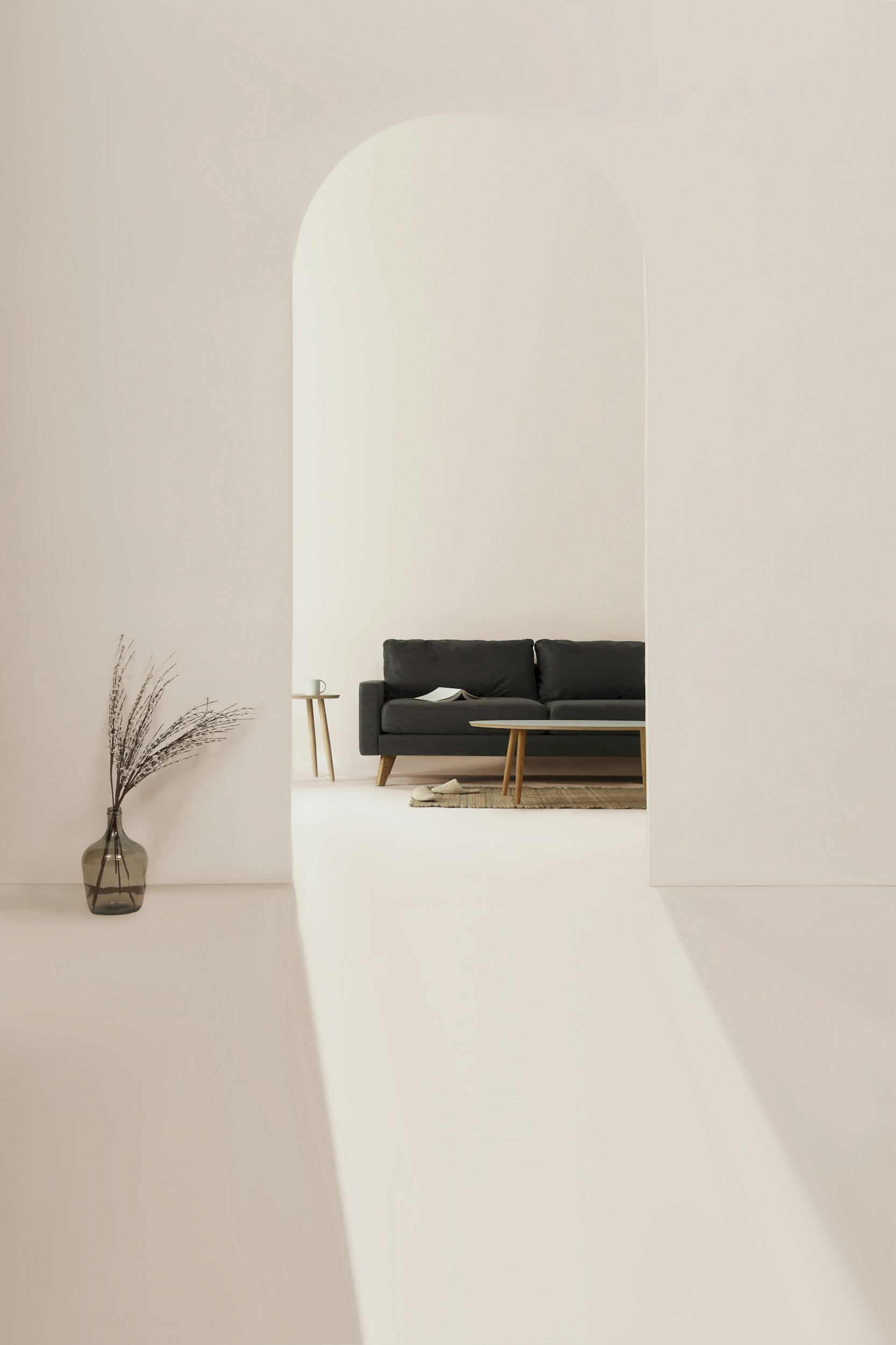 Minimalist living room with a black sofa, wooden coffee table, small side table, and a vase with dried plants, viewed through an arched doorway.