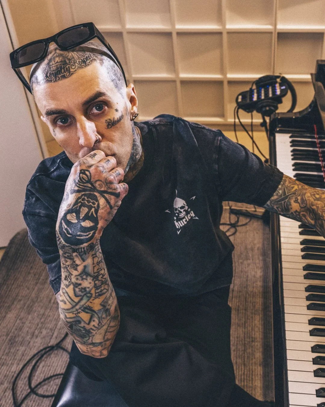 A tattooed man with glasses on his head, sitting at a piano in a recording studio, touching his lips with his tattooed hand.