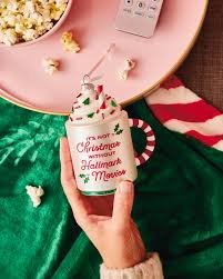 Person holding a mug with Christmas-themed writing and decoration, surrounded by holiday treats and decor on a pink tray, with a green cloth underneath.