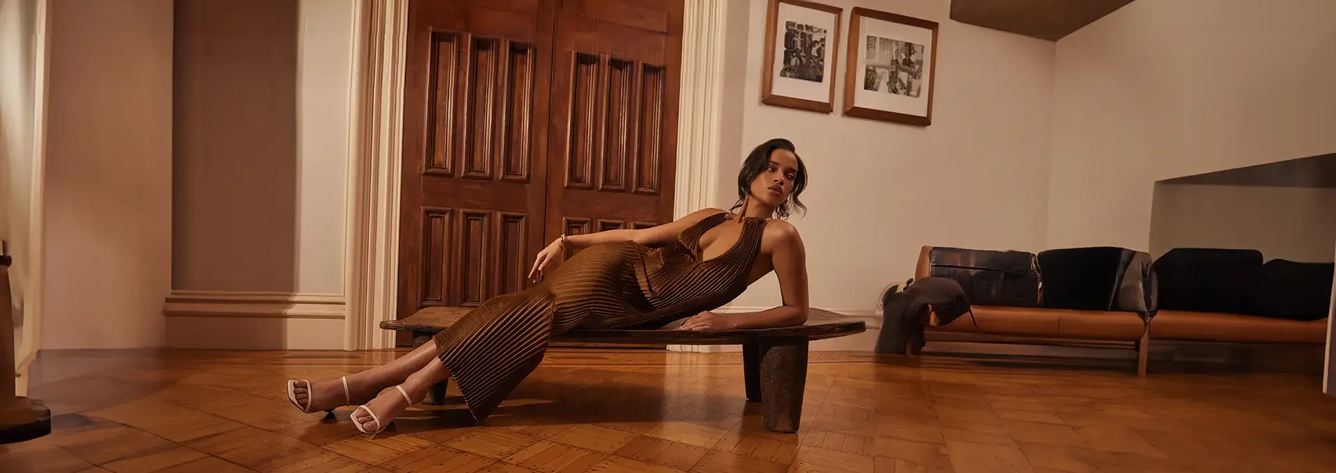 A woman in a brown striped dress and white high heels reclining on a wooden coffee table in a living room, with a doorway, a sofa, and framed pictures on the wall in the background.