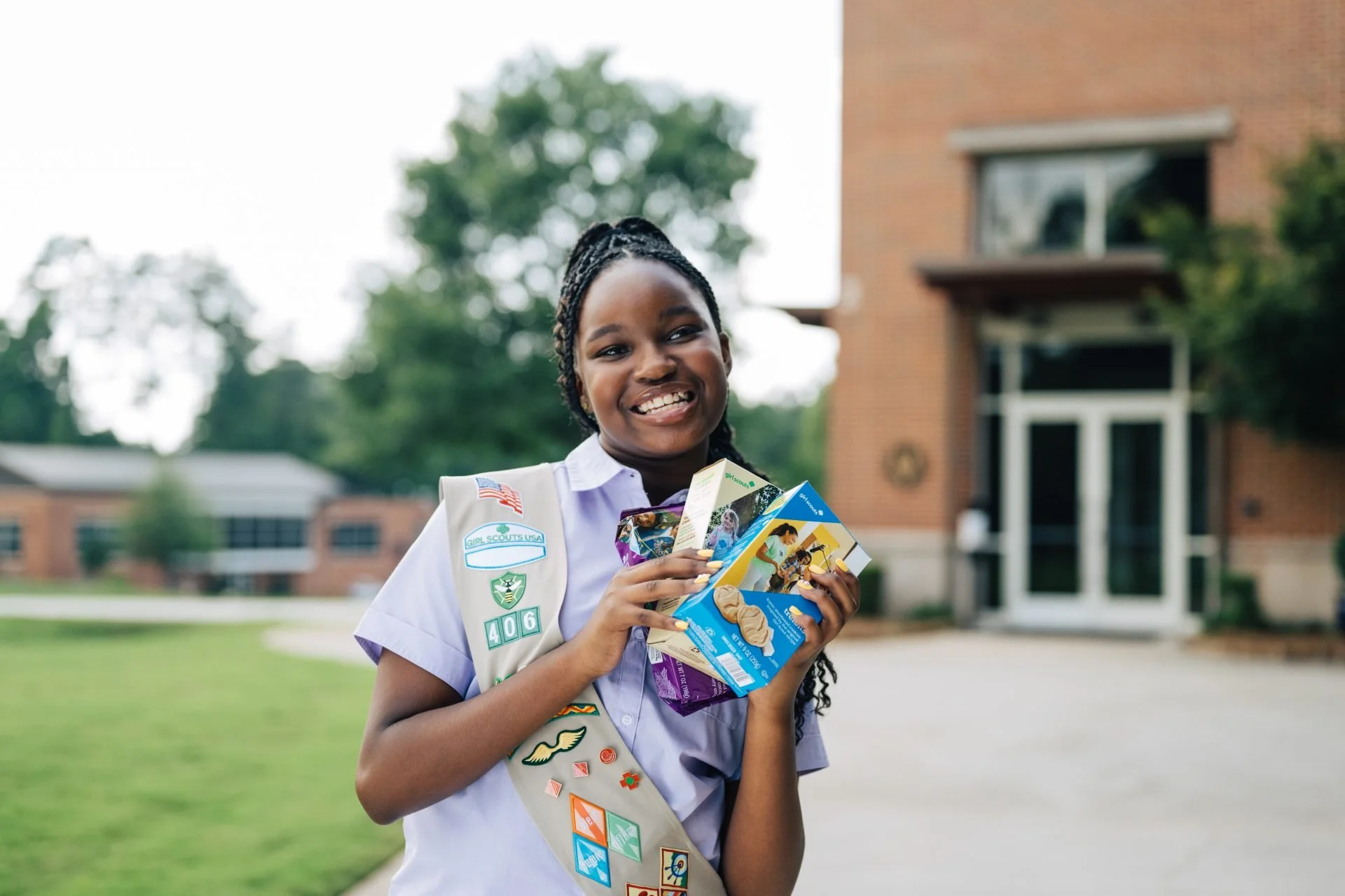 A young girl in a Girl Scouts uniform with badges and a sash, smiling and holding snack boxes outside a brick building and greenery.