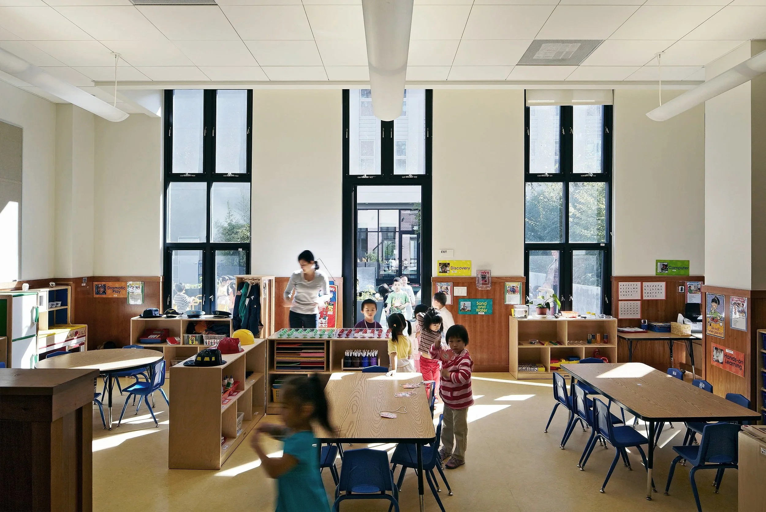 A bright classroom with large windows, children playing and a teacher supervising, organized with tables, chairs, and educational posters.