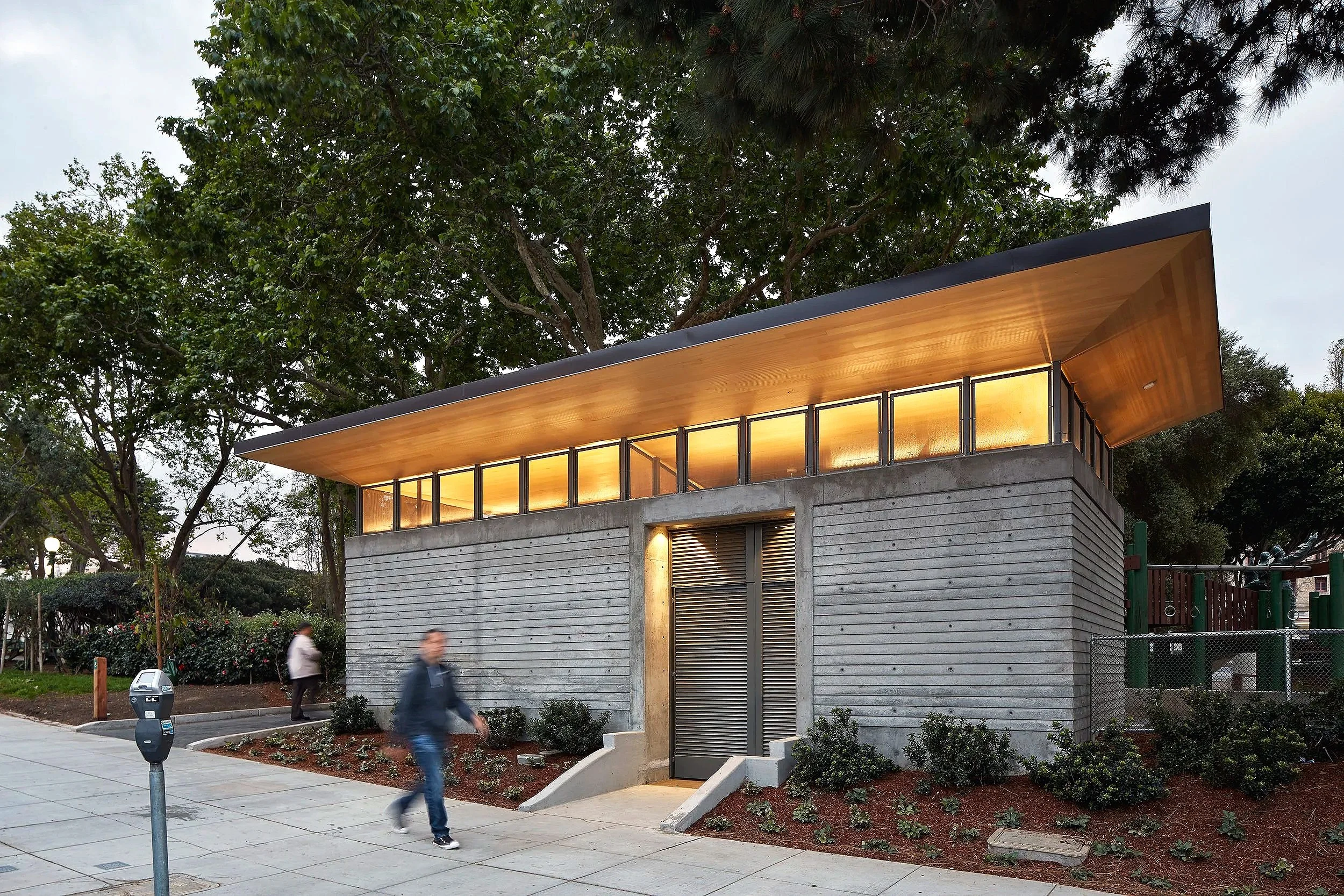 Modern building with concrete walls, a wooden overhanging roof, and a row of illuminated windows at the top. The building is surrounded by trees and bushes, with a sidewalk and a parking meter in the foreground. People are walking past the building.