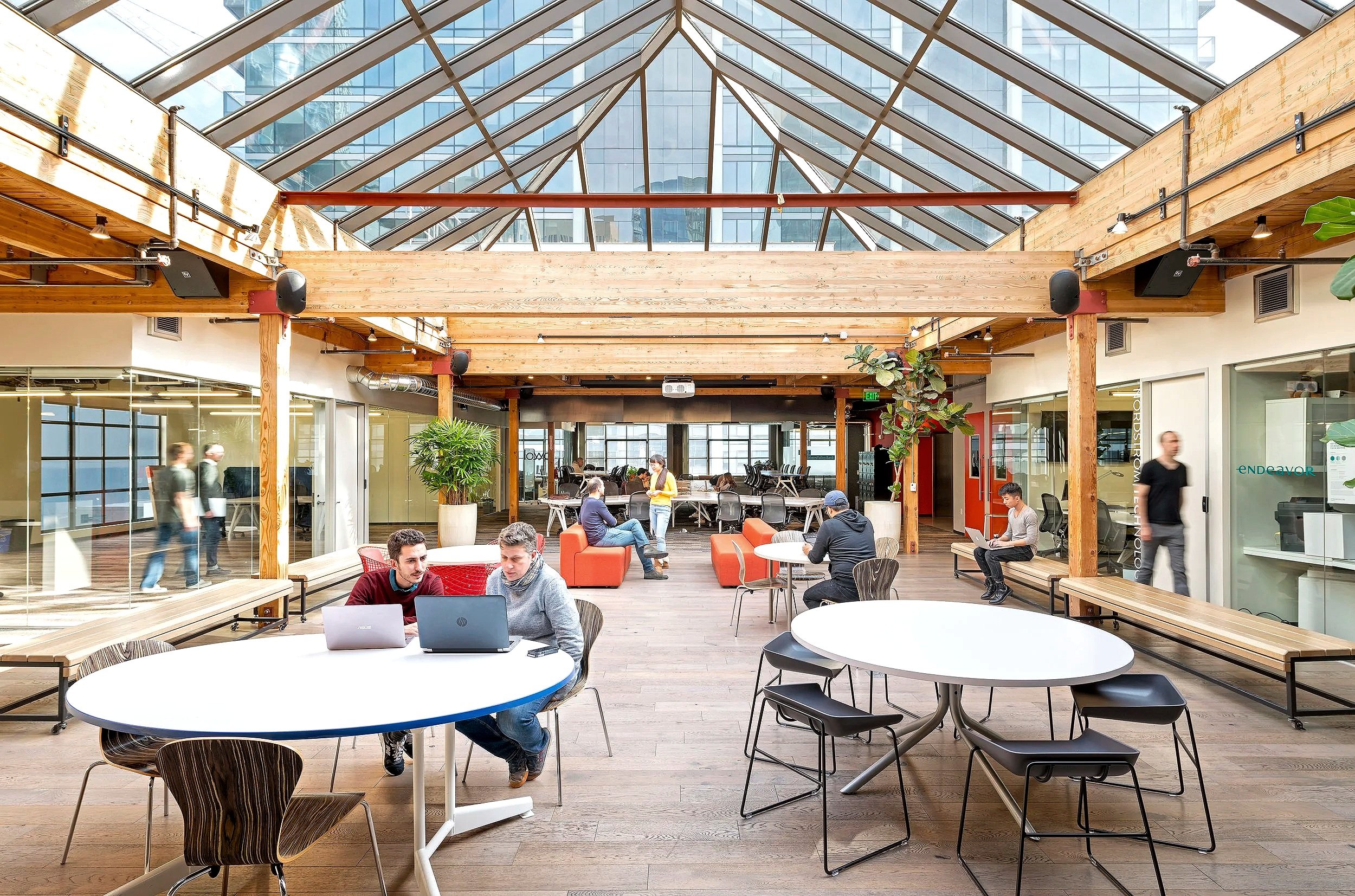 Modern coworking space with wooden beams and lots of natural light from a glass ceiling, people working on laptops and socializing.