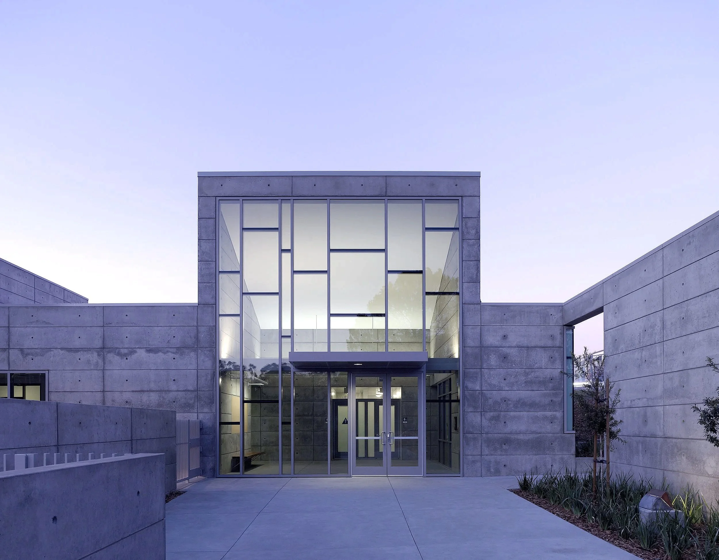 Modern concrete building with large glass windows and doors, surrounded by a paved walkway and landscaped plants.