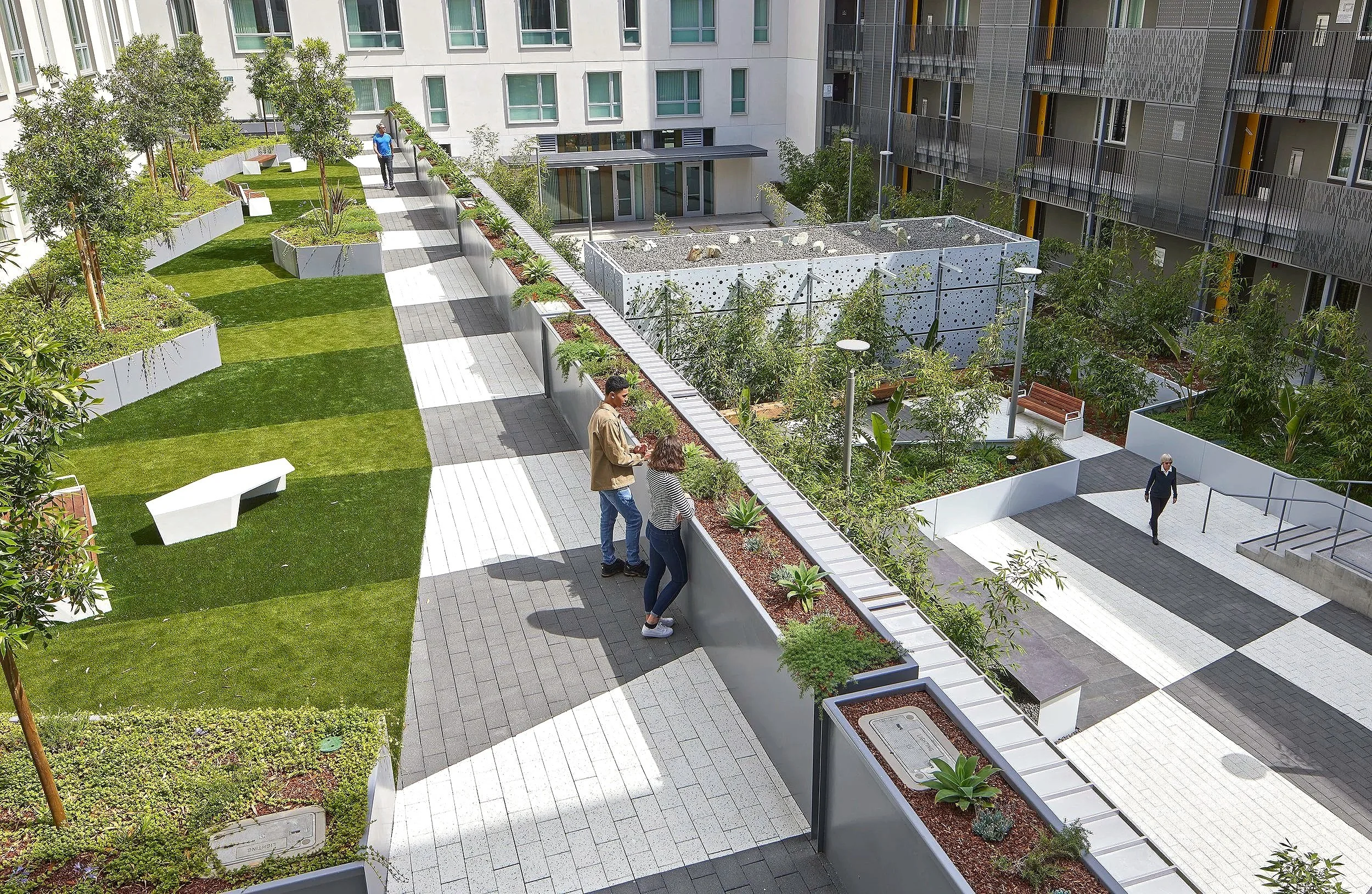 People walking and talking in a modern apartment complex courtyard with greenery, trees, benches, and pathways.