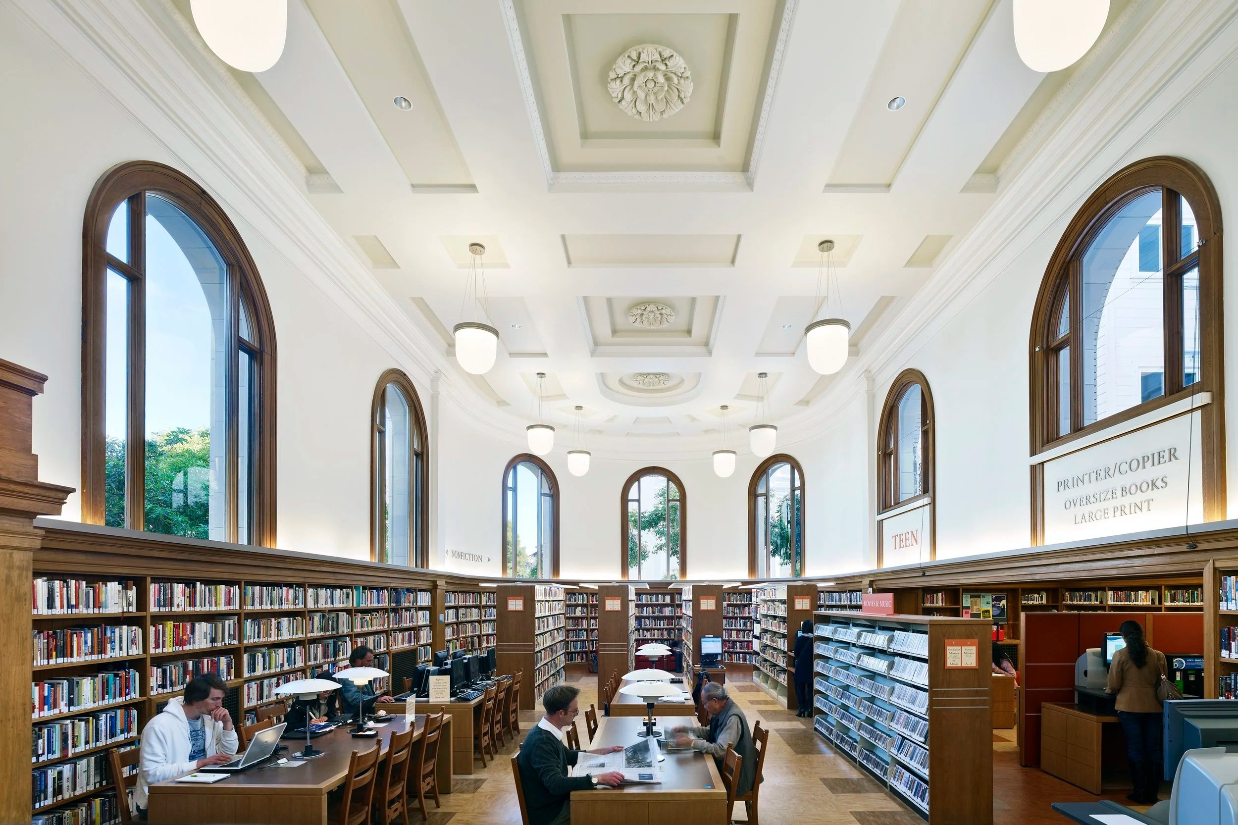 Interior of a library with high ceilings, large arched windows, bookshelves, and people working at tables.