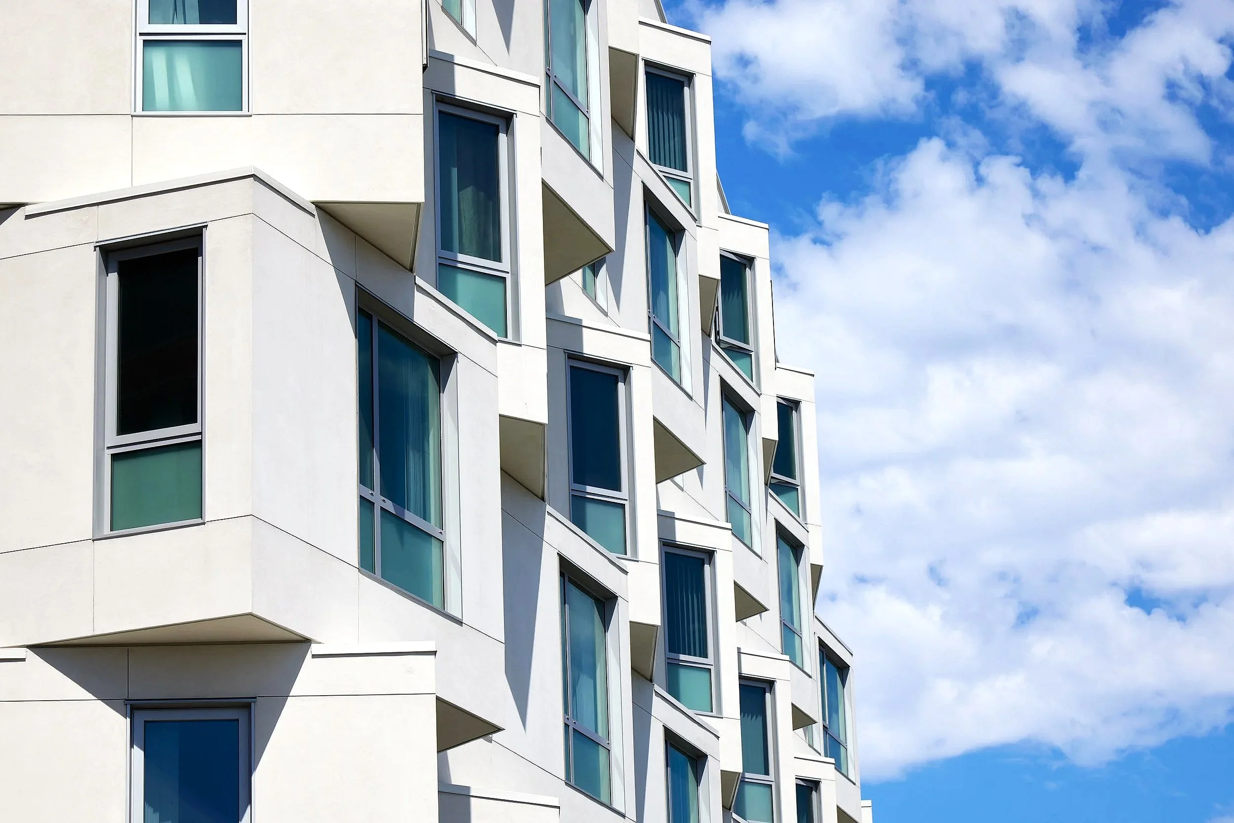 Modern white apartment building with large windows and small balconies, against a blue sky with clouds.
